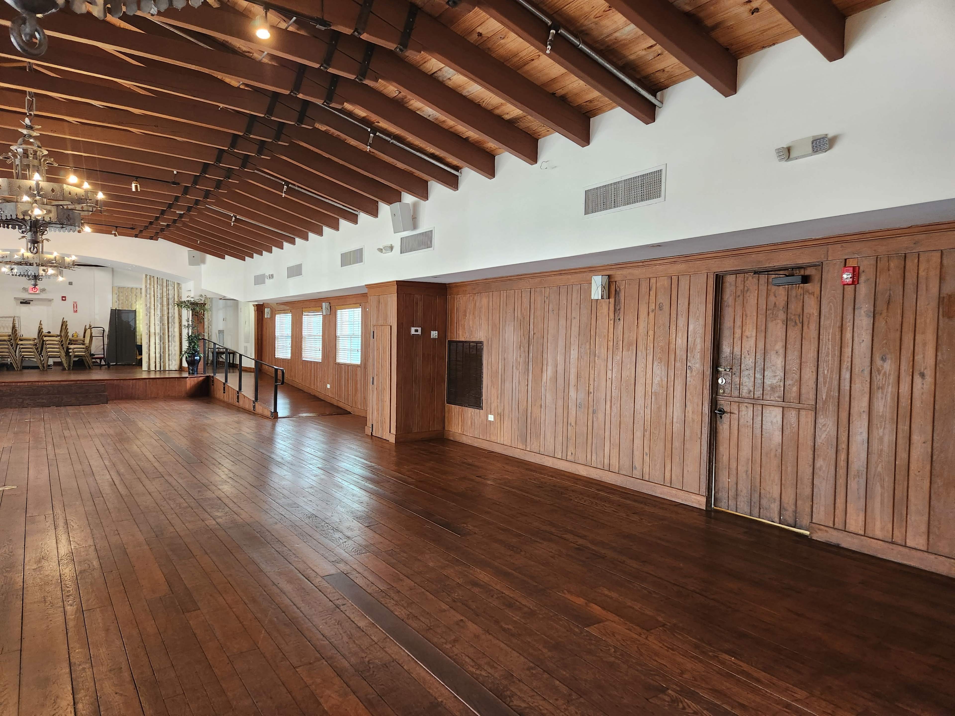 A spacious interior room with wooden beams on the ceiling, polished wooden floors, and a set of folding chairs arranged against one wall.
