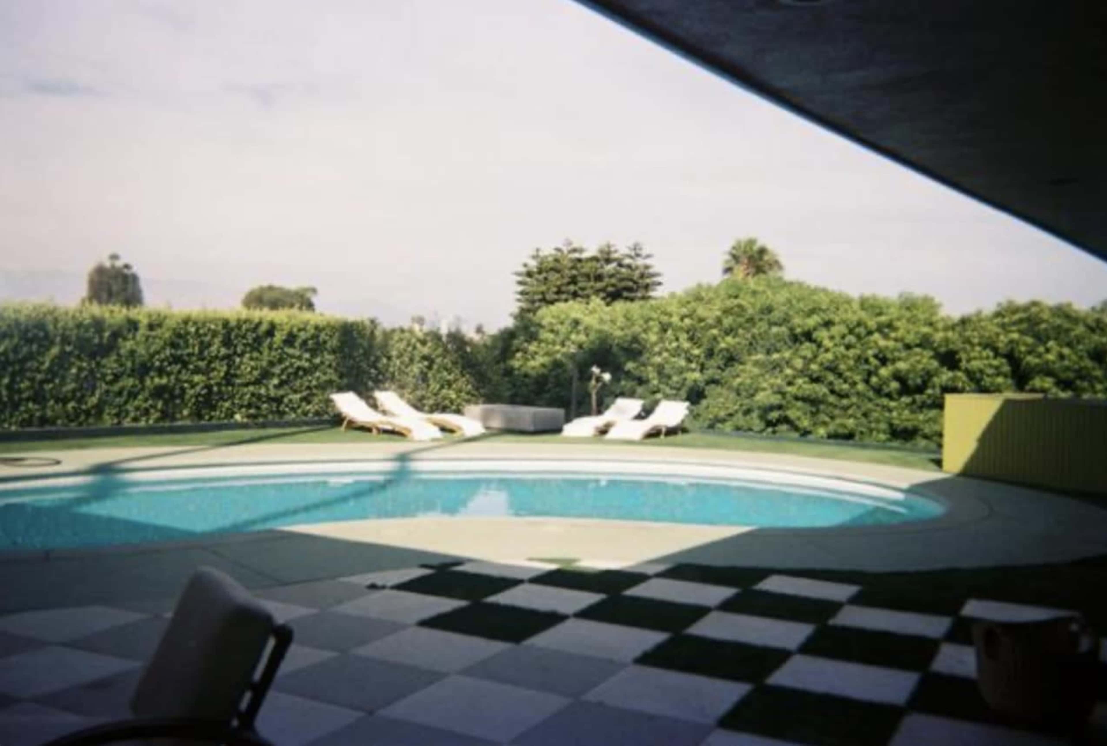 The image shows a swimming pool surrounded by green foliage and lounge chairs on a patio.