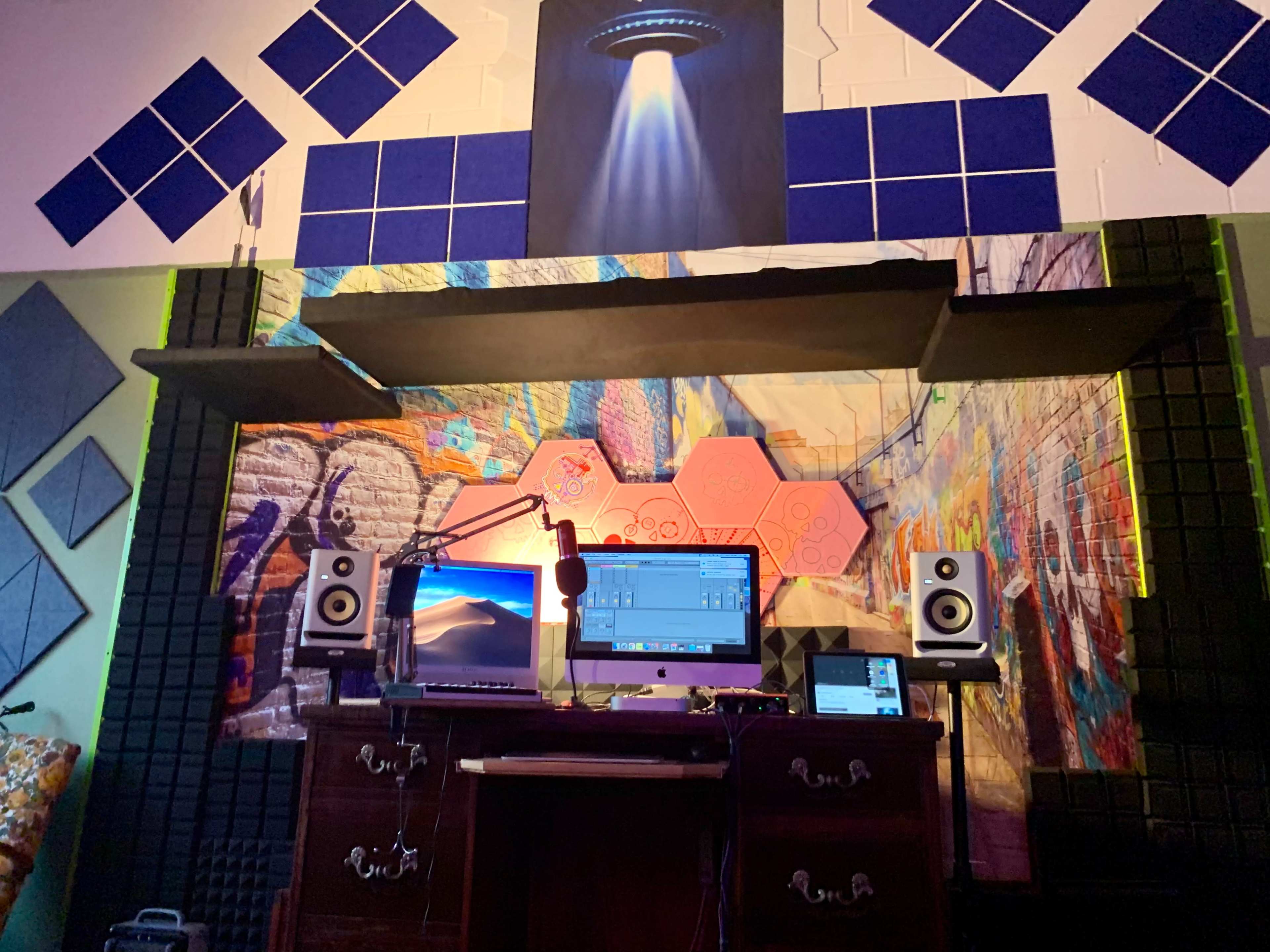 A music production setup featuring a desk with two computer monitors, a microphone, speakers, and colorful wall art in a studio environment.