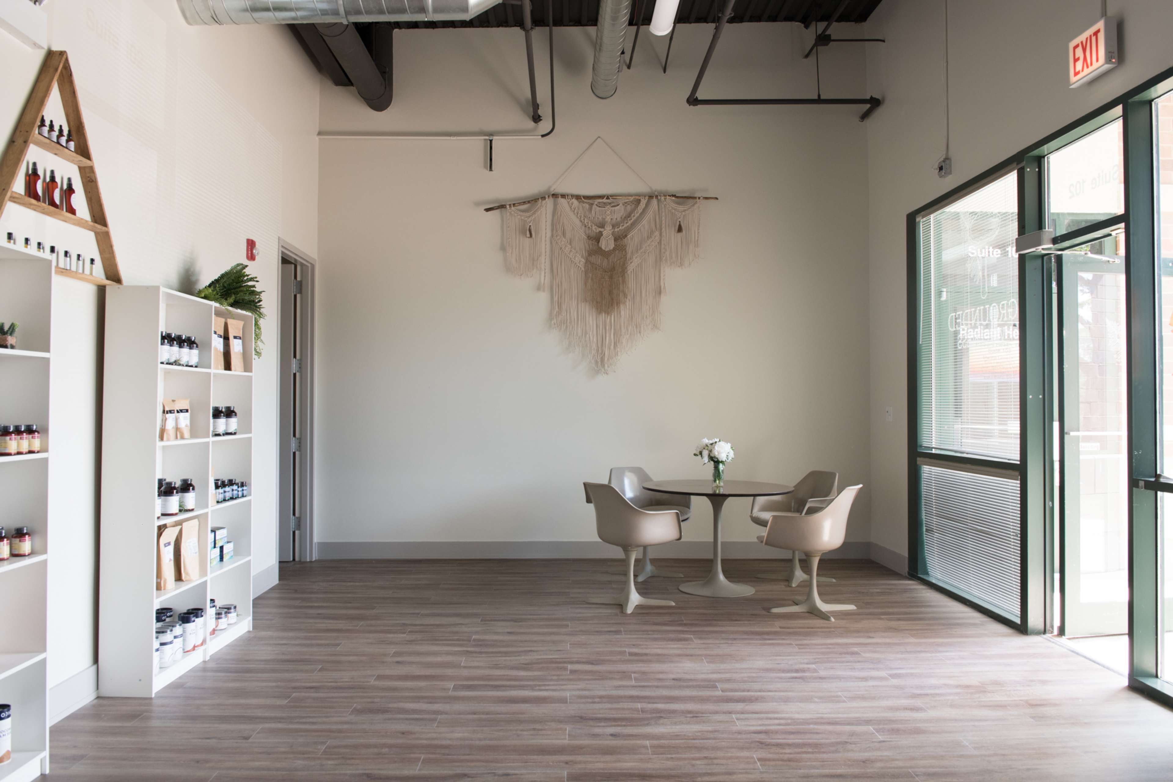A minimalist interior of a retail space featuring a seating area with a table and two chairs, a wall hanging, and shelving units filled with products.