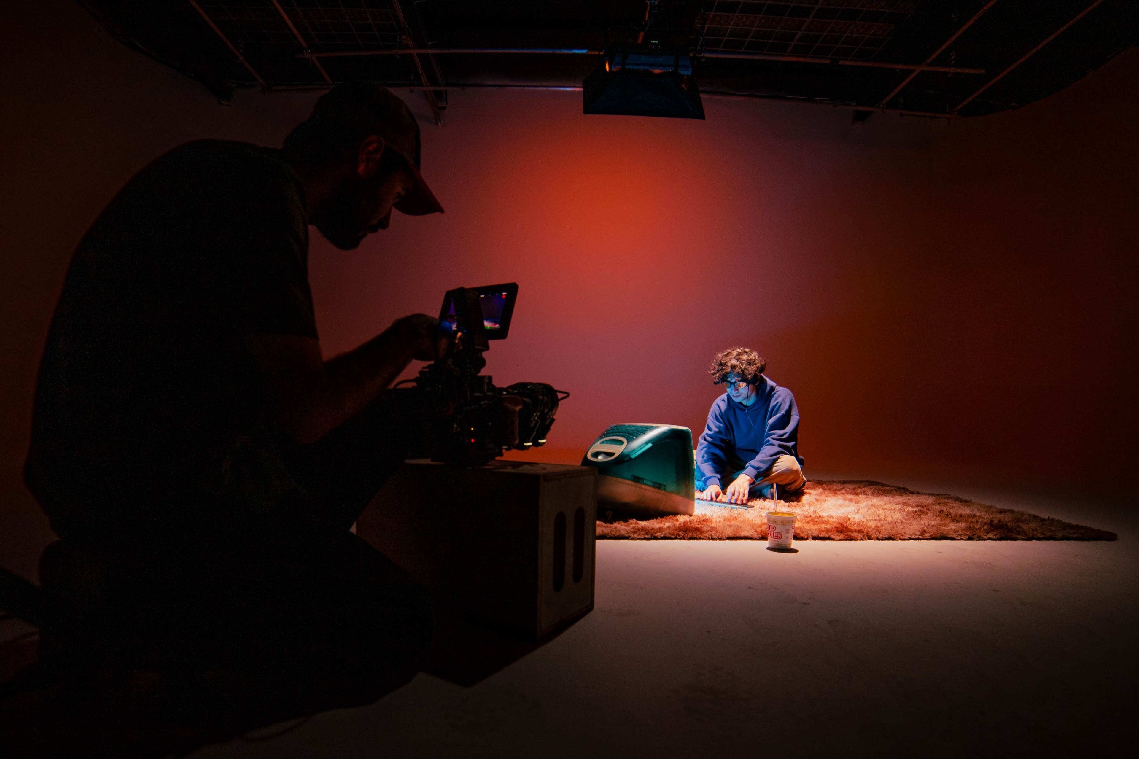 A person sits on a rug in a dimly lit studio, while another person operates a camera, capturing the scene.