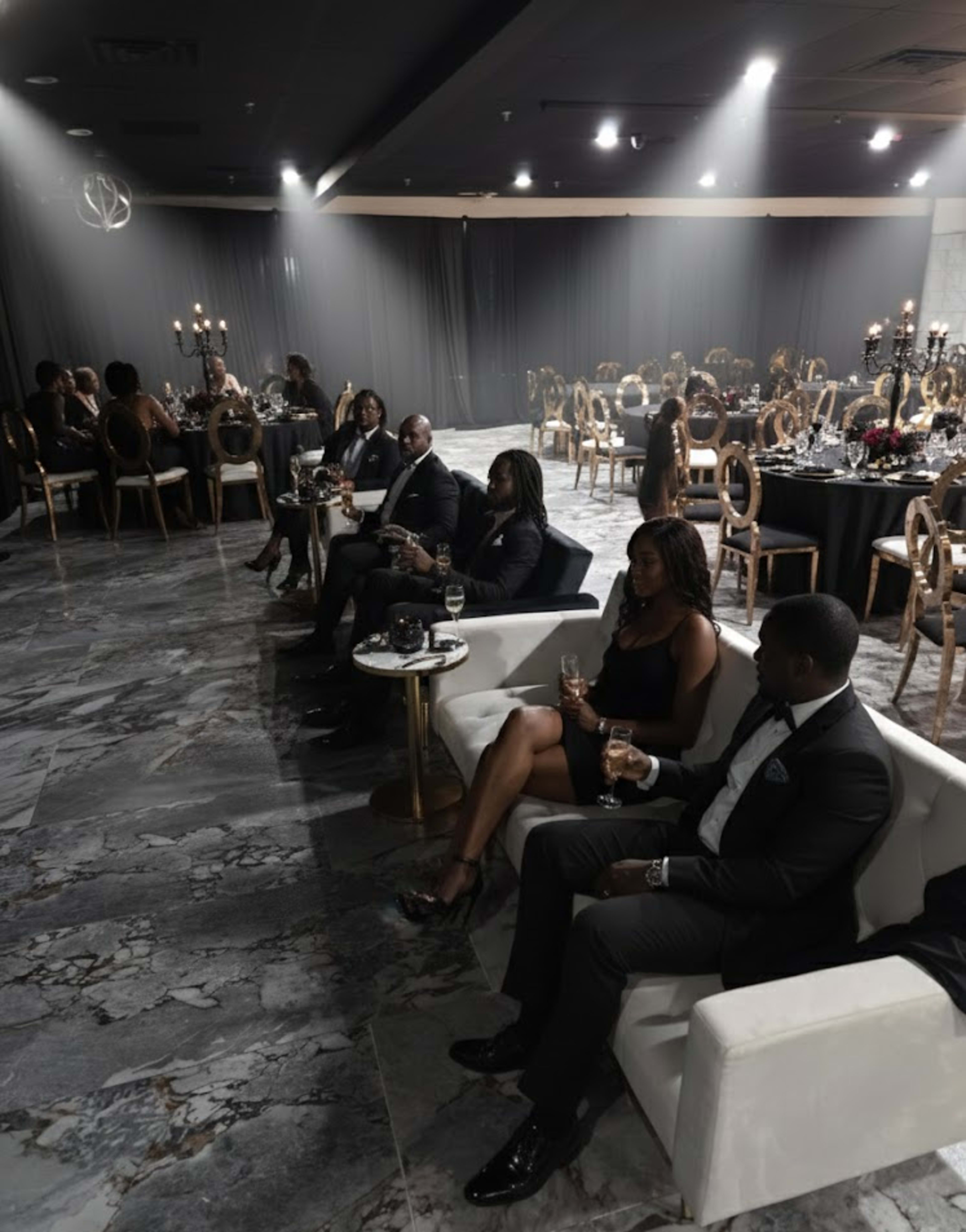 A group of elegantly dressed individuals sits on a white sofa in a dimly lit banquet hall adorned with tables set for a formal event.