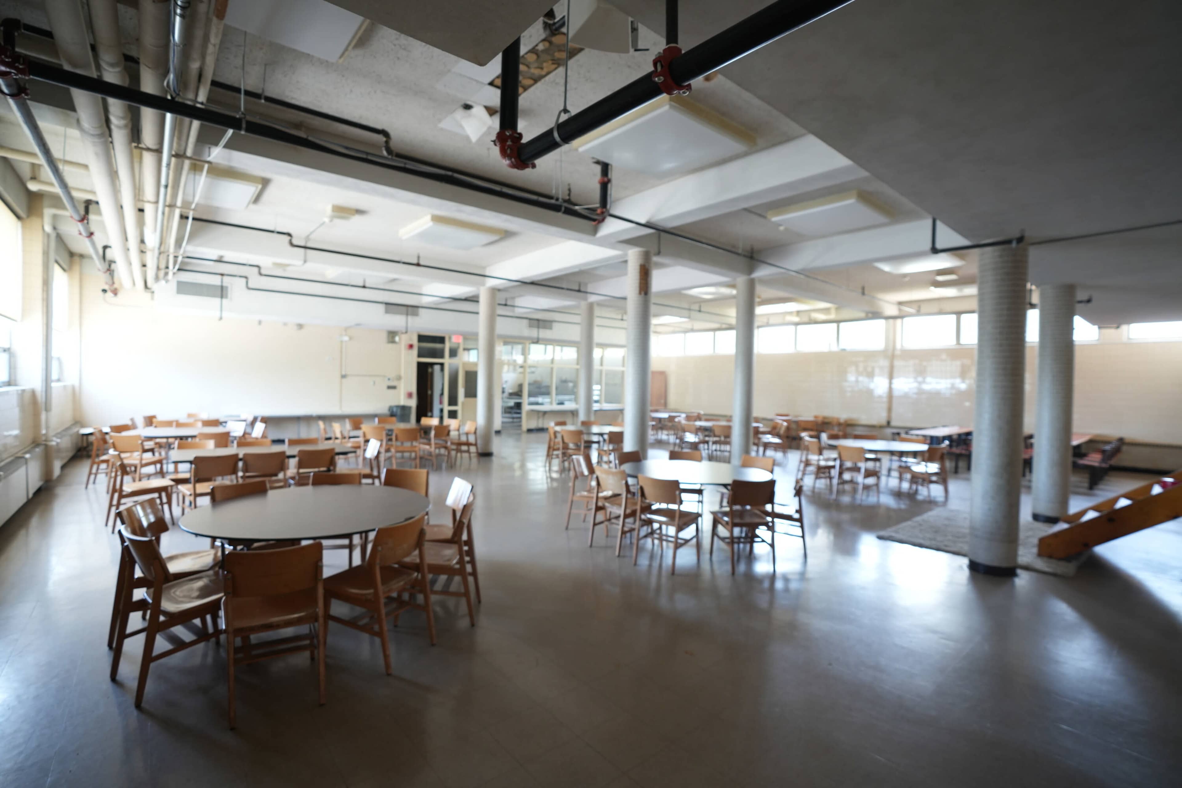 The image shows a spacious cafeteria with circular tables and wooden chairs arranged throughout the room.