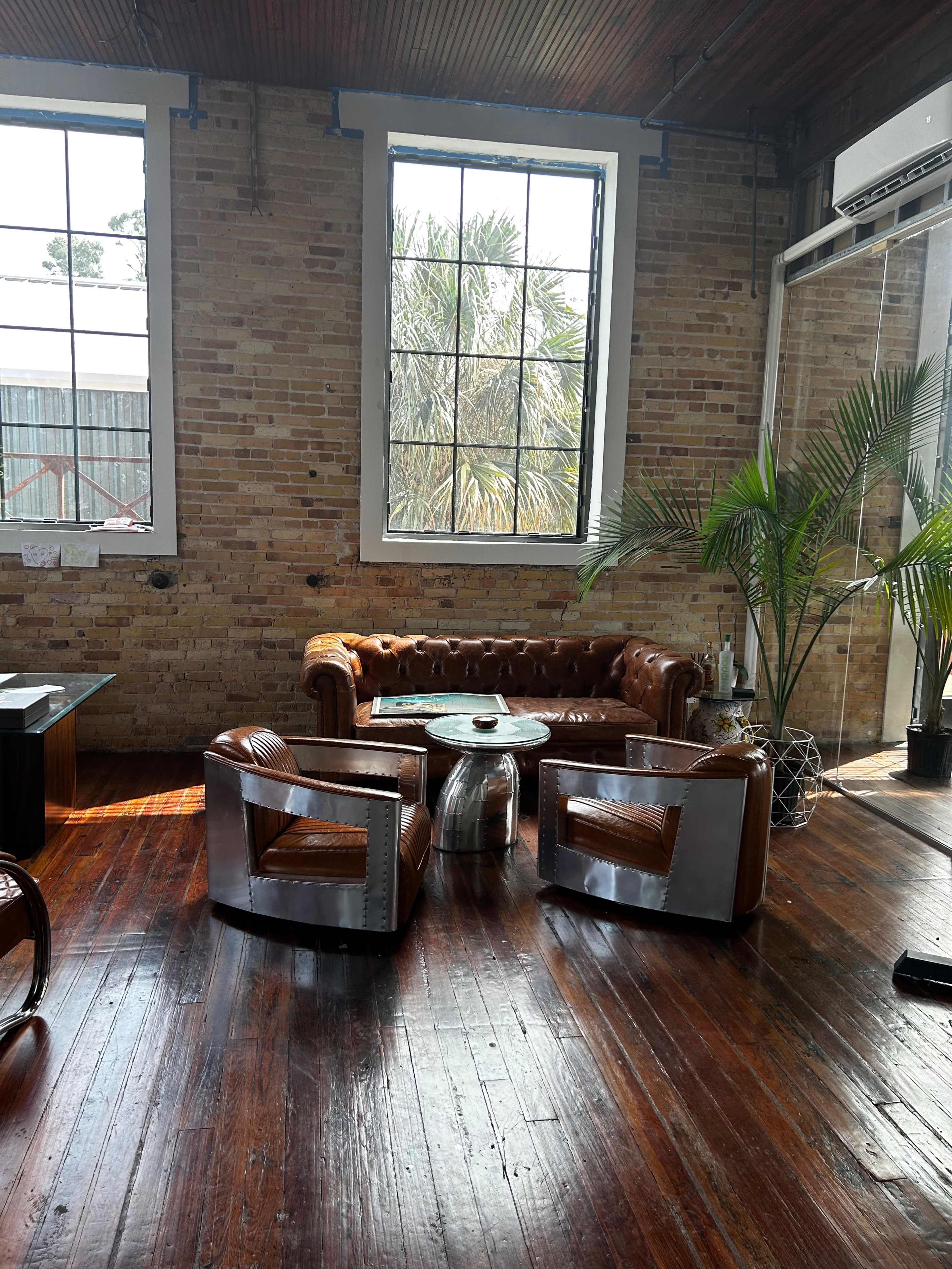 The scene features a leather chesterfield sofa and two contemporary chairs surrounded by large windows and a palm plant in a room with exposed brick walls and polished wooden floors.