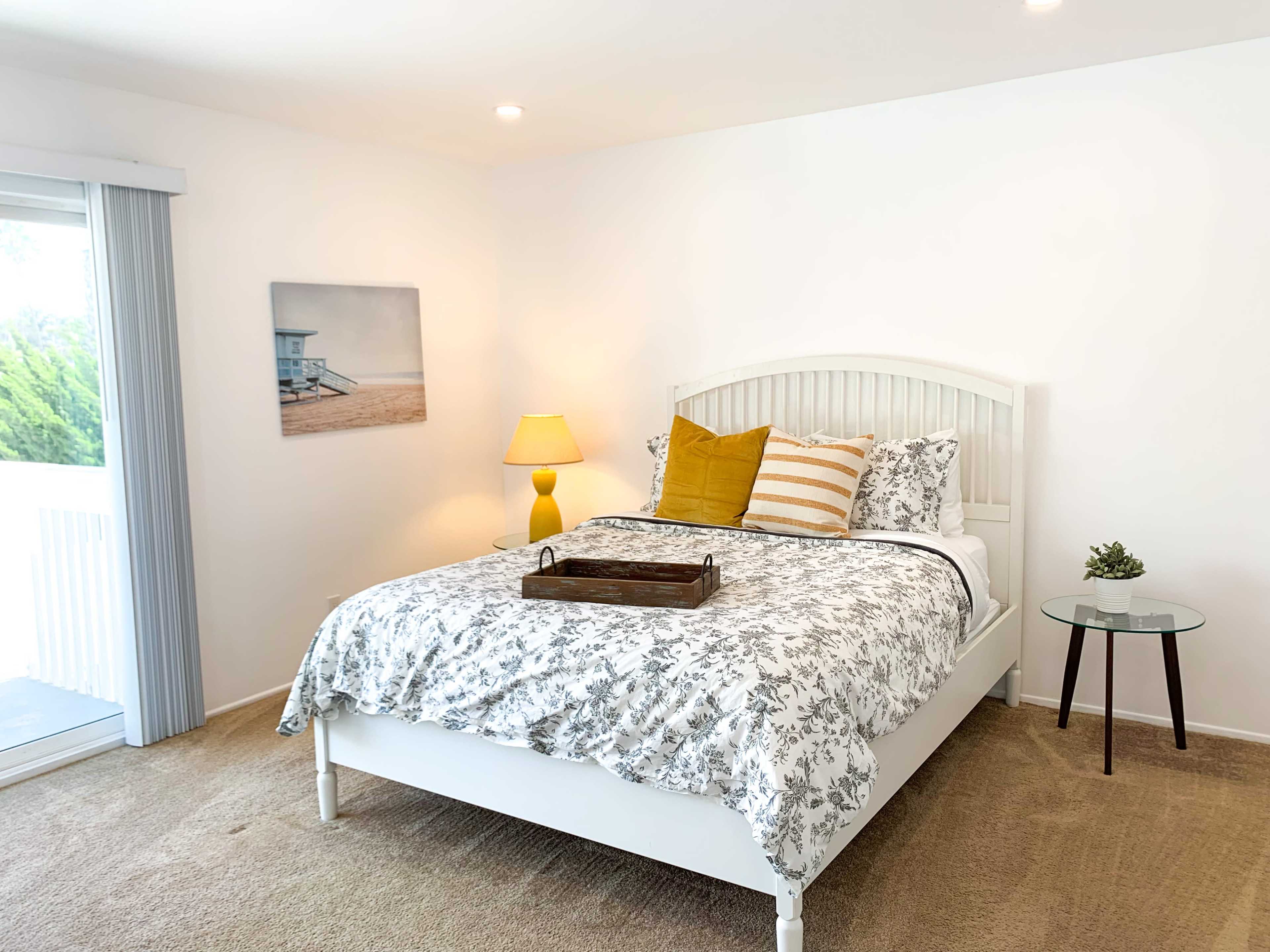 A neatly arranged bedroom features a white bed with decorative pillows, a wooden tray on the bed, a lamp, and a side table next to a sliding glass door.
