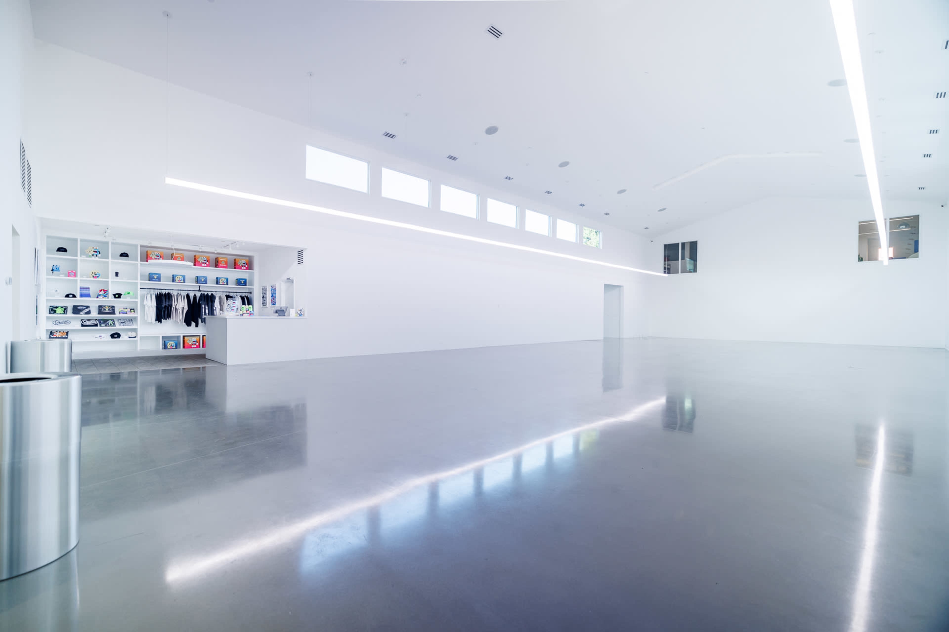 The image shows a spacious, brightly lit interior with a polished concrete floor and minimalist design, featuring shelving on one wall displaying various items.