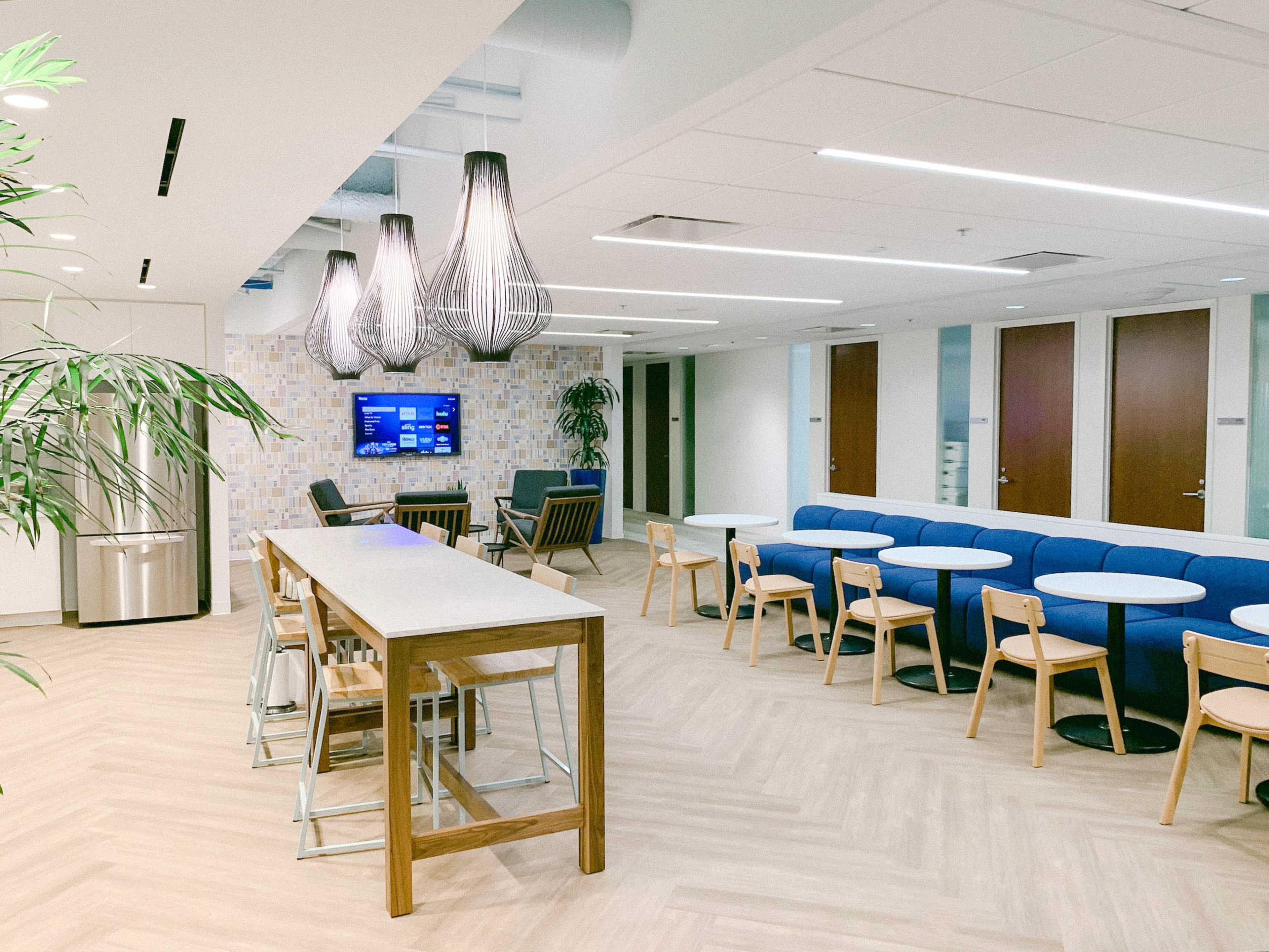 A modern office break room features wooden tables, blue seating, and pendant lighting, alongside a wall-mounted television and indoor plants.
