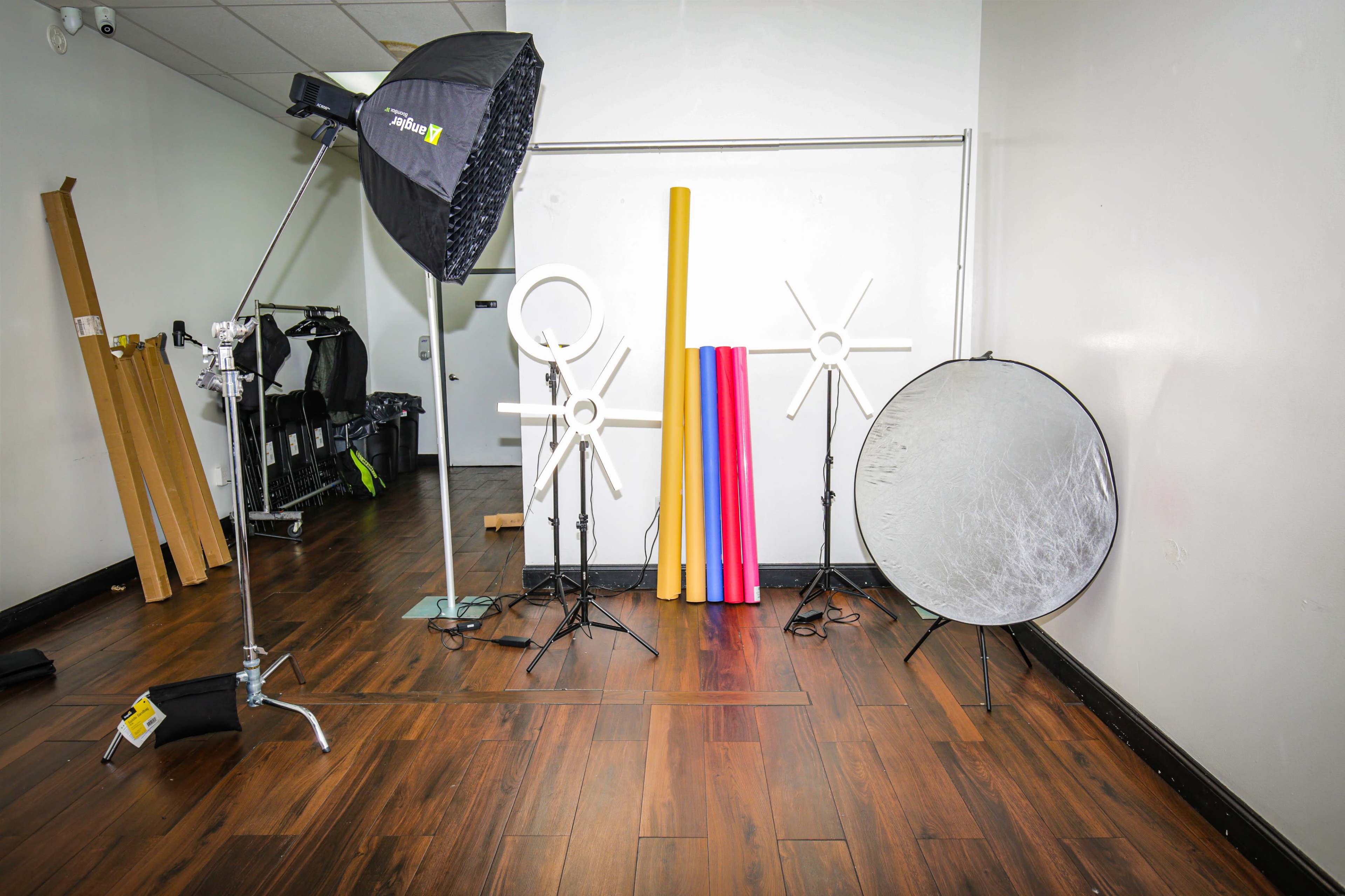 The image shows a photography studio setup featuring a softbox light, several background rolls, and a reflector on a wooden floor.
