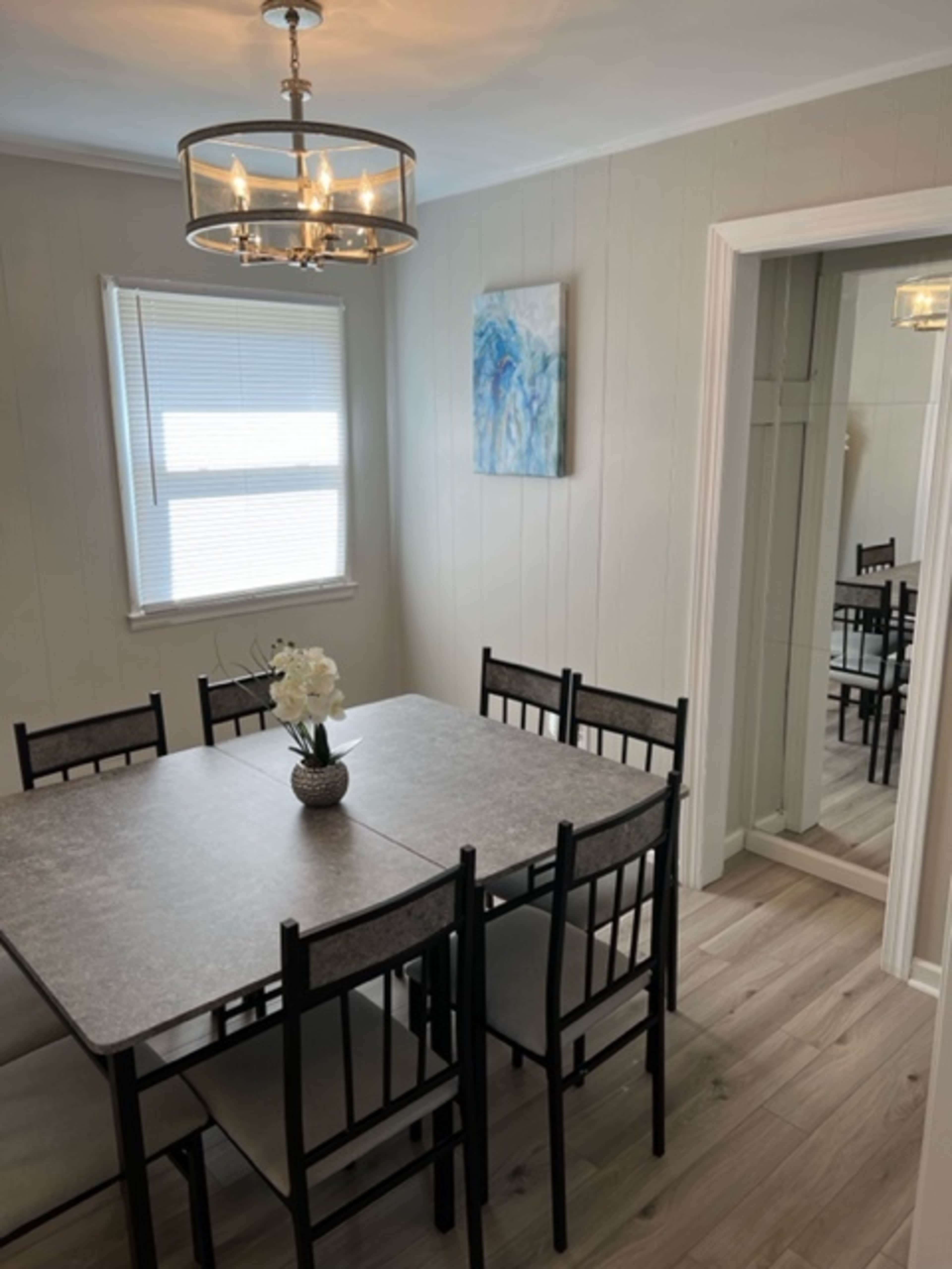 A dining room features a rectangular table surrounded by six black chairs, with a light fixture above and a window letting in natural light.