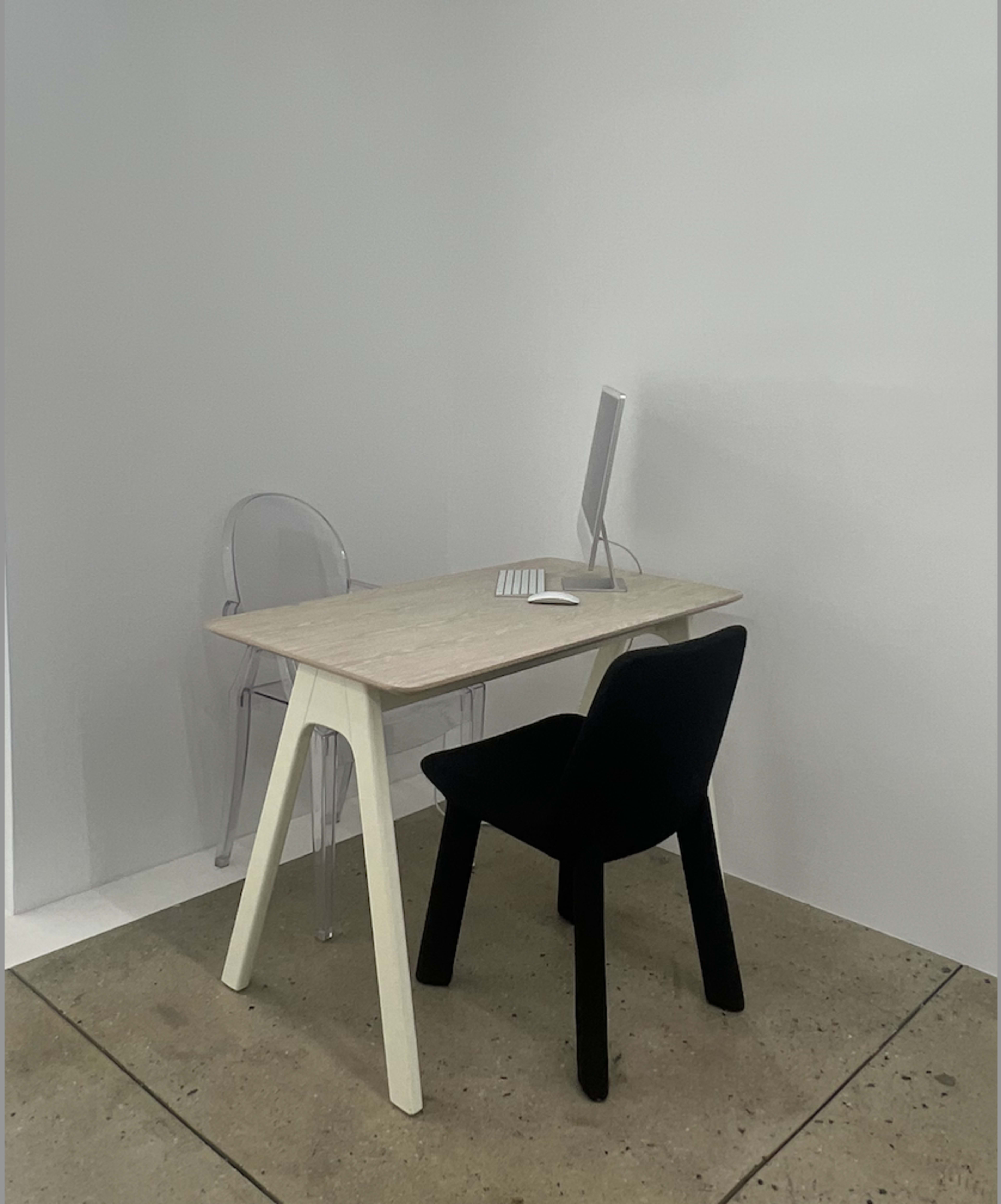 A minimalist workspace features a light-colored table with a computer and a black chair, set against a plain wall.