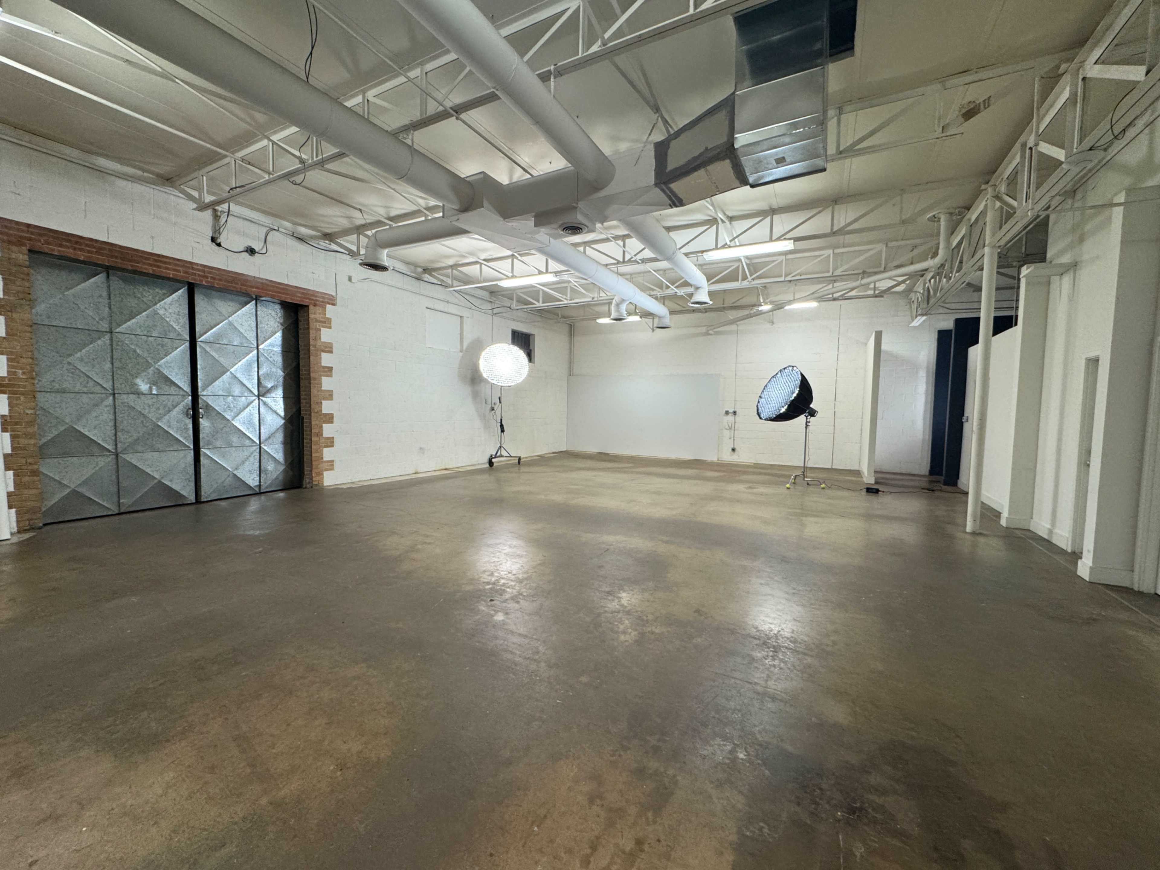 The image shows an empty, industrial-style room with high ceilings, two large light softboxes, and a pair of metallic doors on one side.