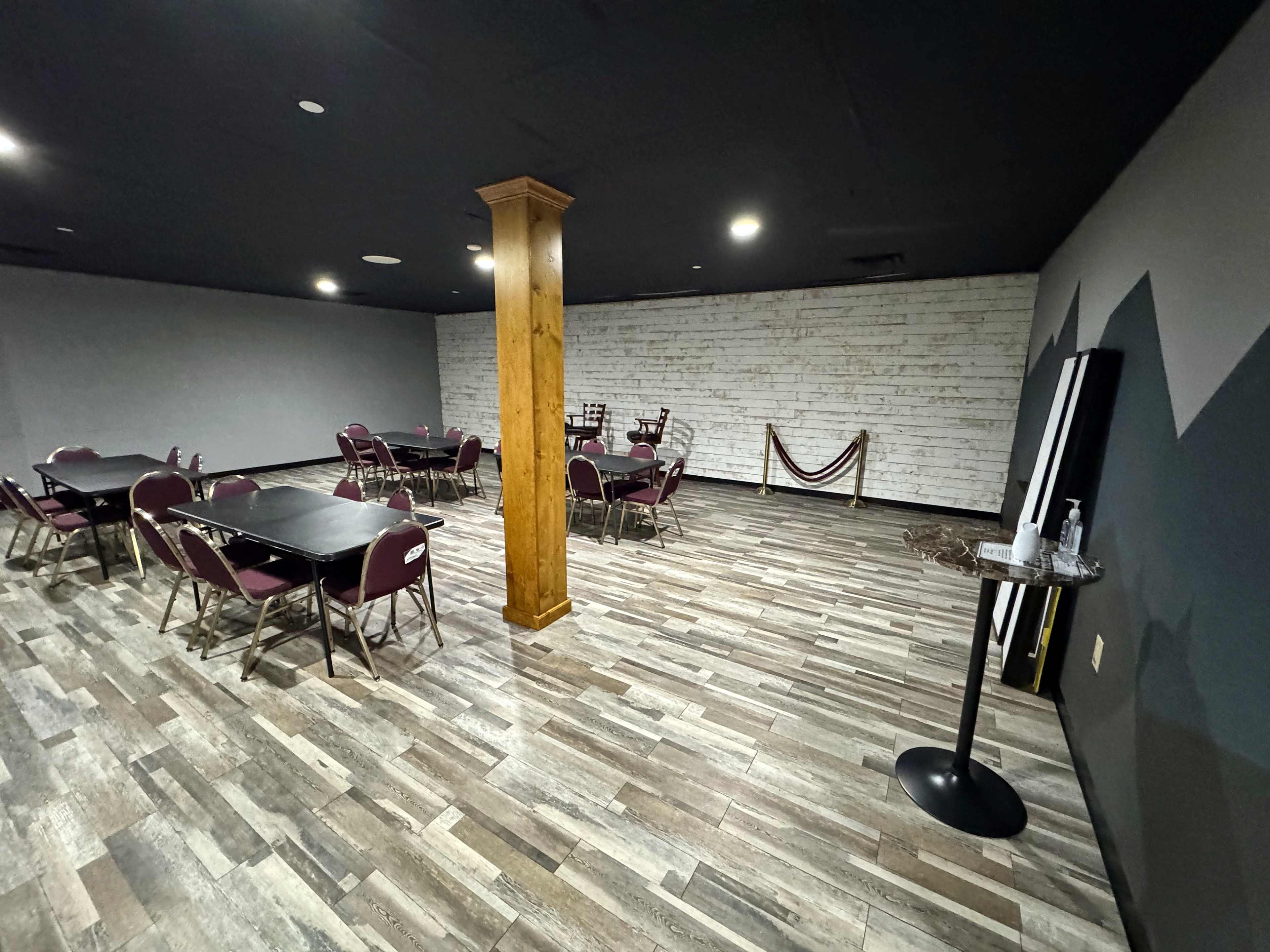 The image shows an empty, dimly lit event space featuring several round tables and chairs, with wooden flooring and a decorative column in the center.