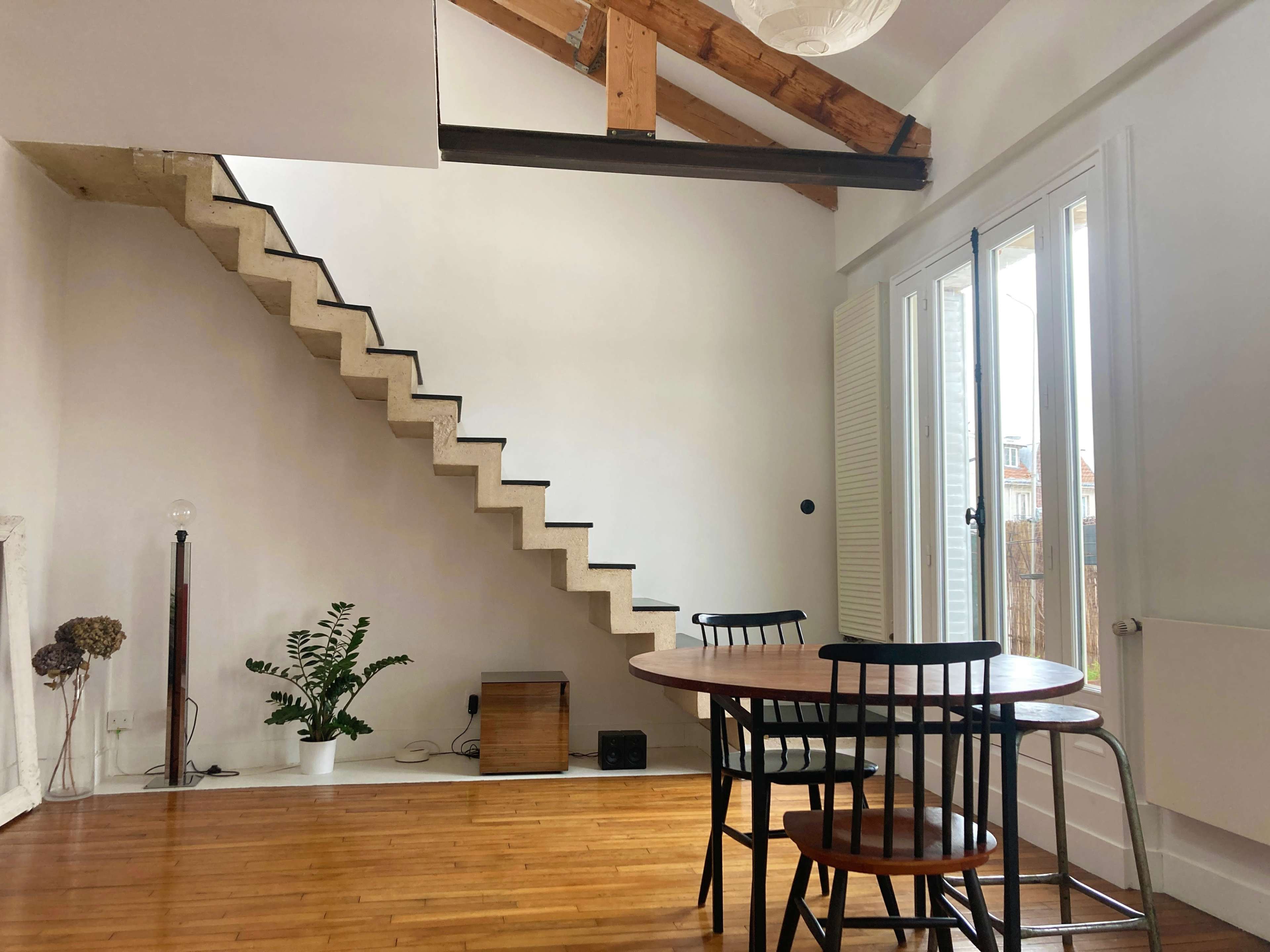 A modern interior features a round dining table with black chairs, a staircase leading to an upper level, and large windows providing natural light.