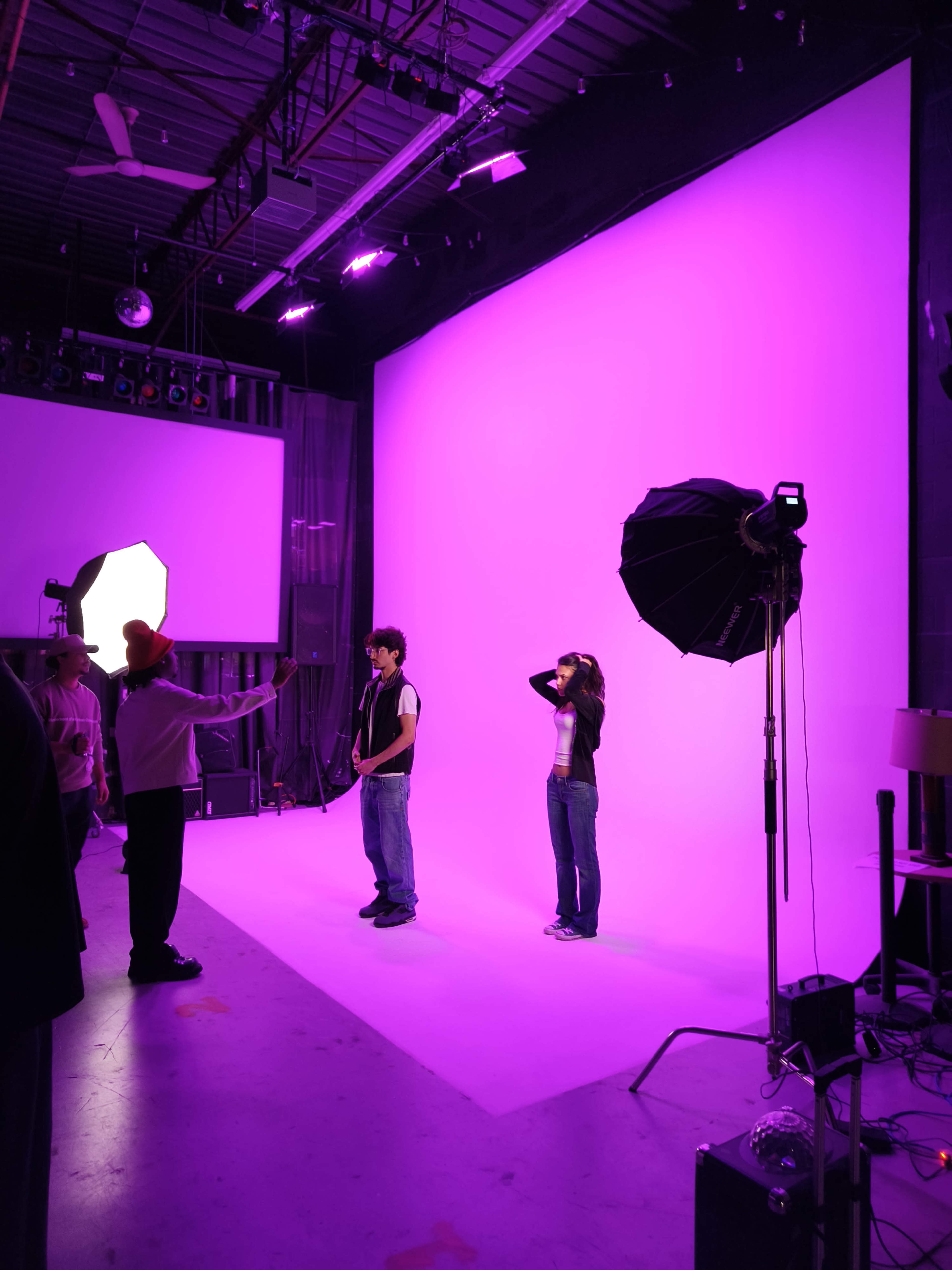 Two models stand in front of a bright purple backdrop in a photography studio, while a photographer directs them.