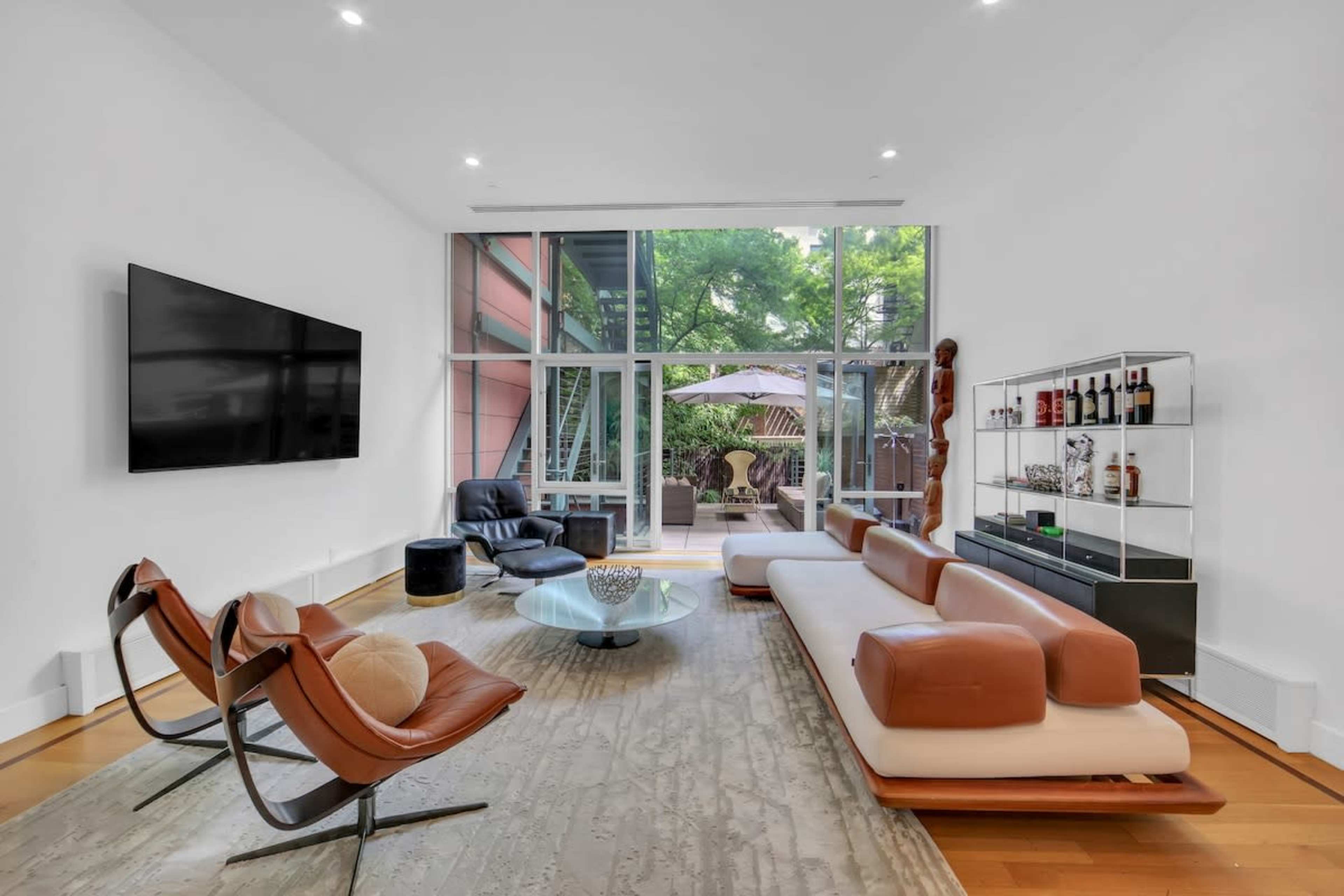 The living room features a large window that opens to a patio, with modern furniture including a brown leather sectional sofa and black lounge chair set around a glass coffee table.