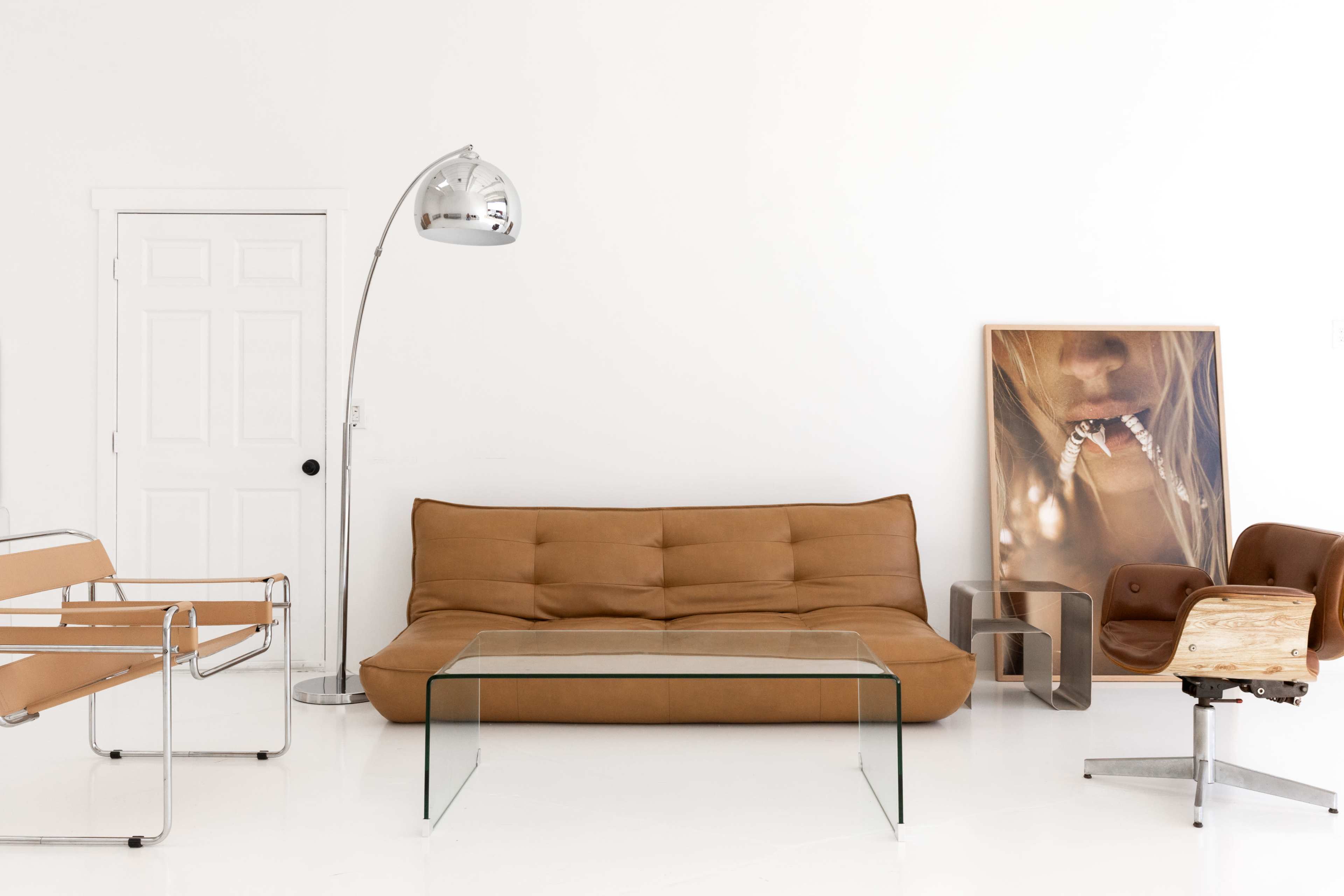 The image shows a minimalist living room featuring a brown sofa, a glass coffee table, two modern chairs, and a large framed artwork on a white wall.