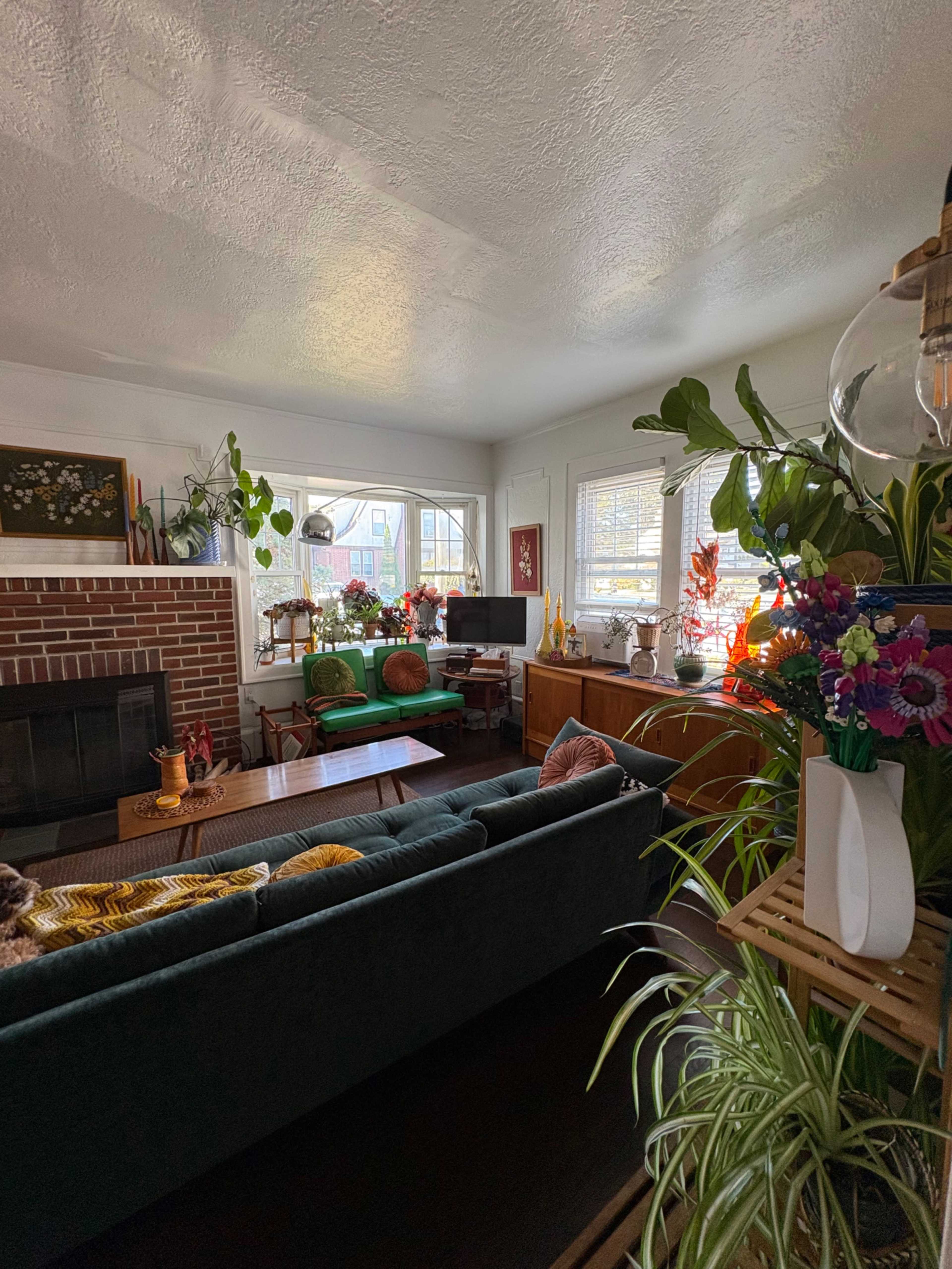 The image shows a brightly lit living room featuring a dark green sofa, a brick fireplace, potted plants, and various pieces of furniture including a wooden cabinet and green armchairs.