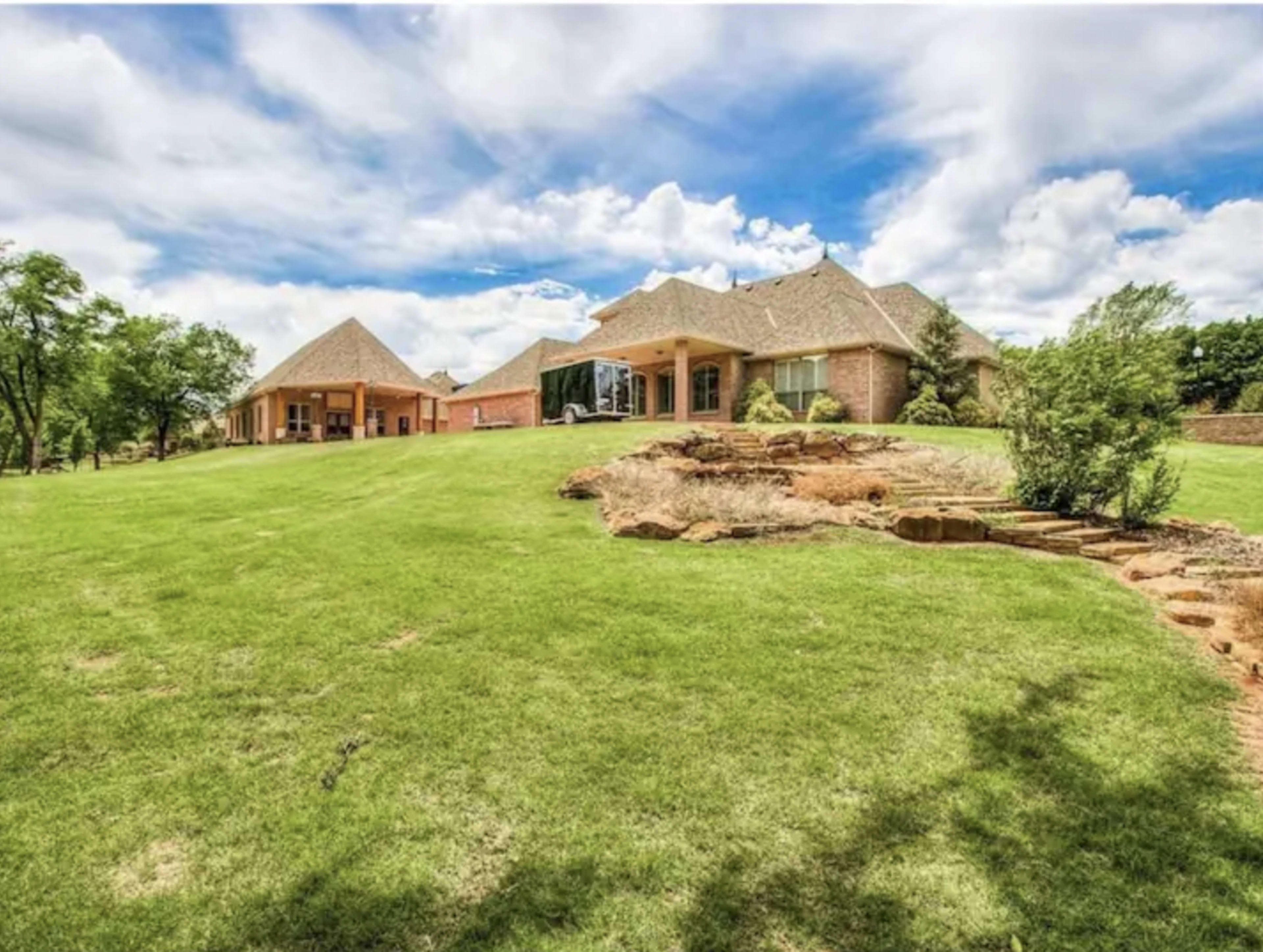 A large residential property features a brick house and a separate building on a lush green lawn under a partly cloudy sky.