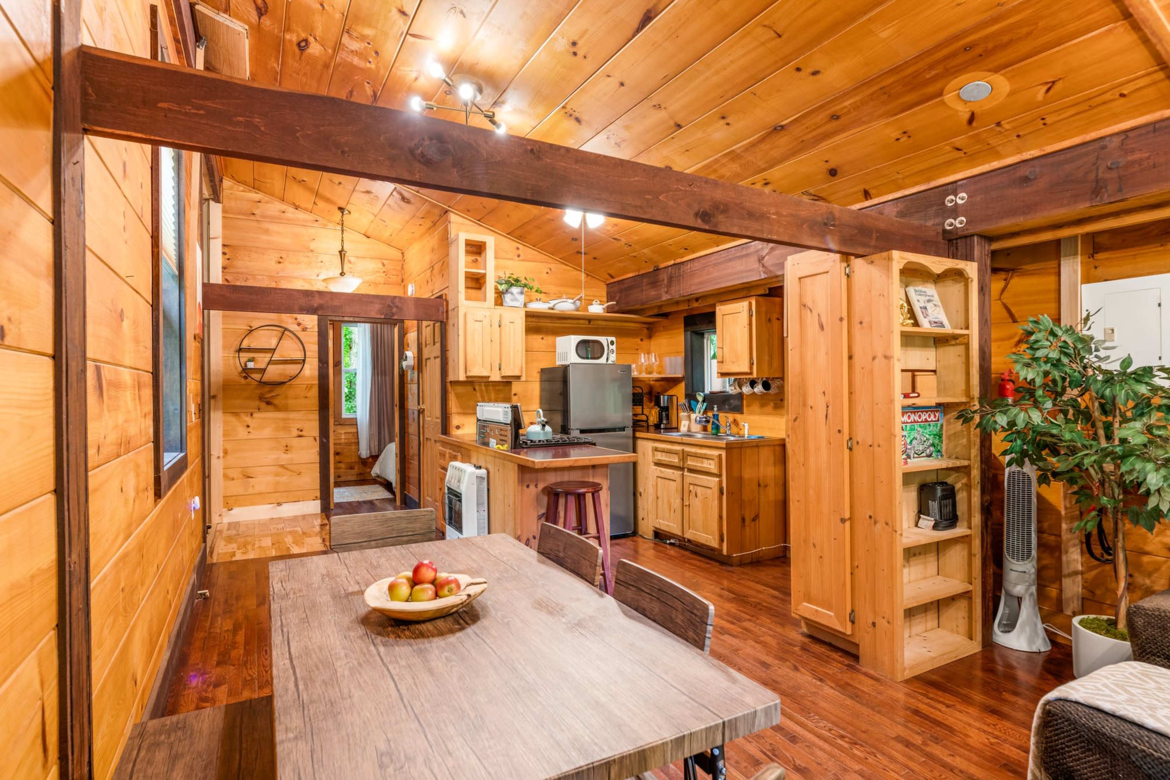 A wooden interior of a cabin featuring a kitchen area, a dining table with fruit, and living space with wooden beams.