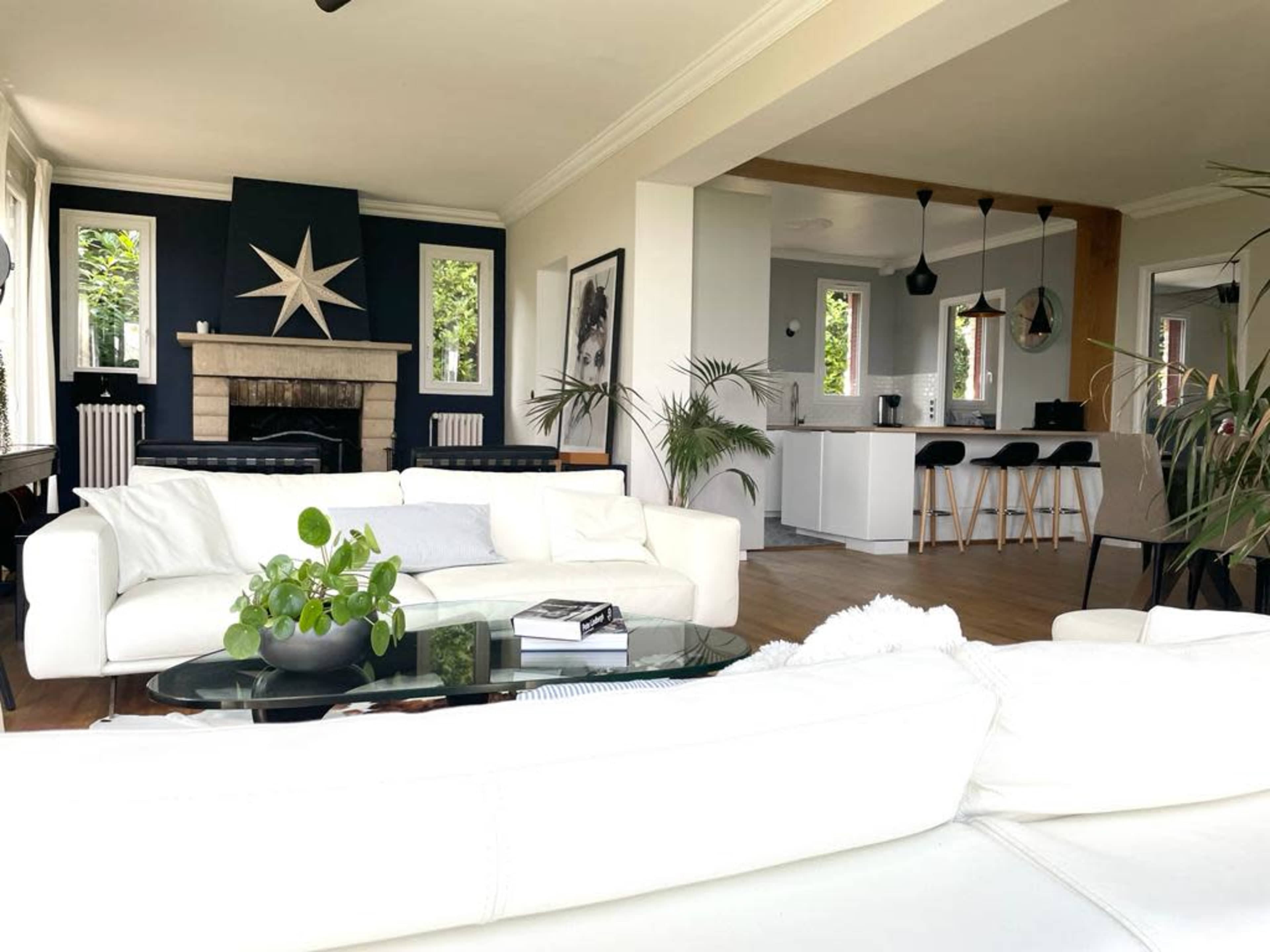 A modern living room features a white sectional sofa, a glass coffee table with a plant, and a stylish fireplace, leading to an open kitchen with a minimalist design.