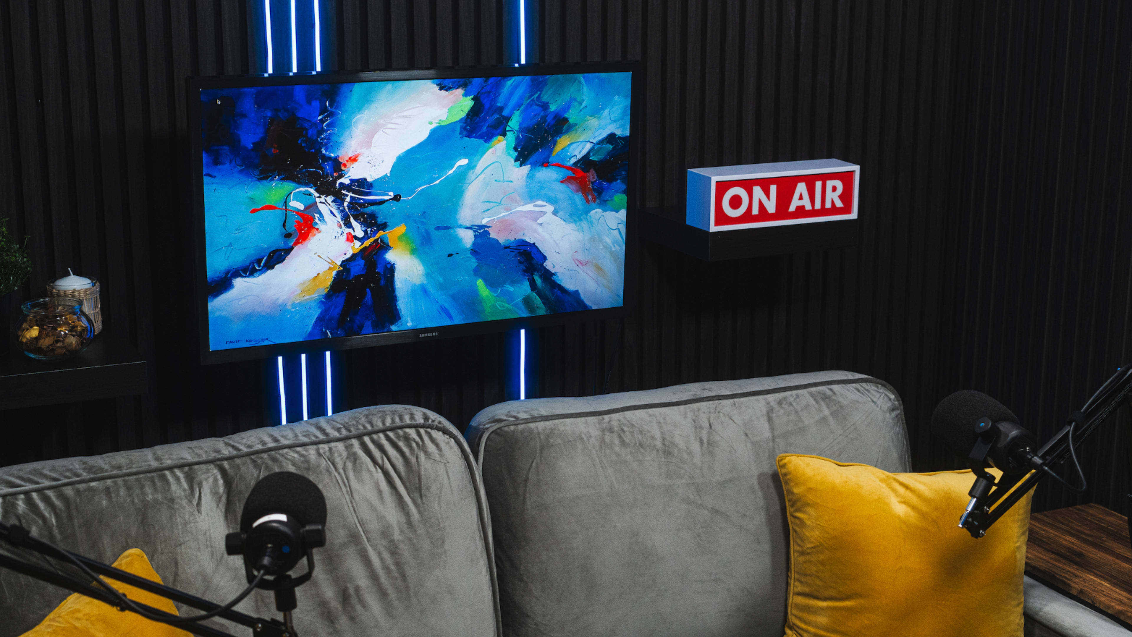 A painting with abstract colors hangs on a wall next to an "ON AIR" sign, above a gray sofa with yellow cushions and two microphones set for a recording.