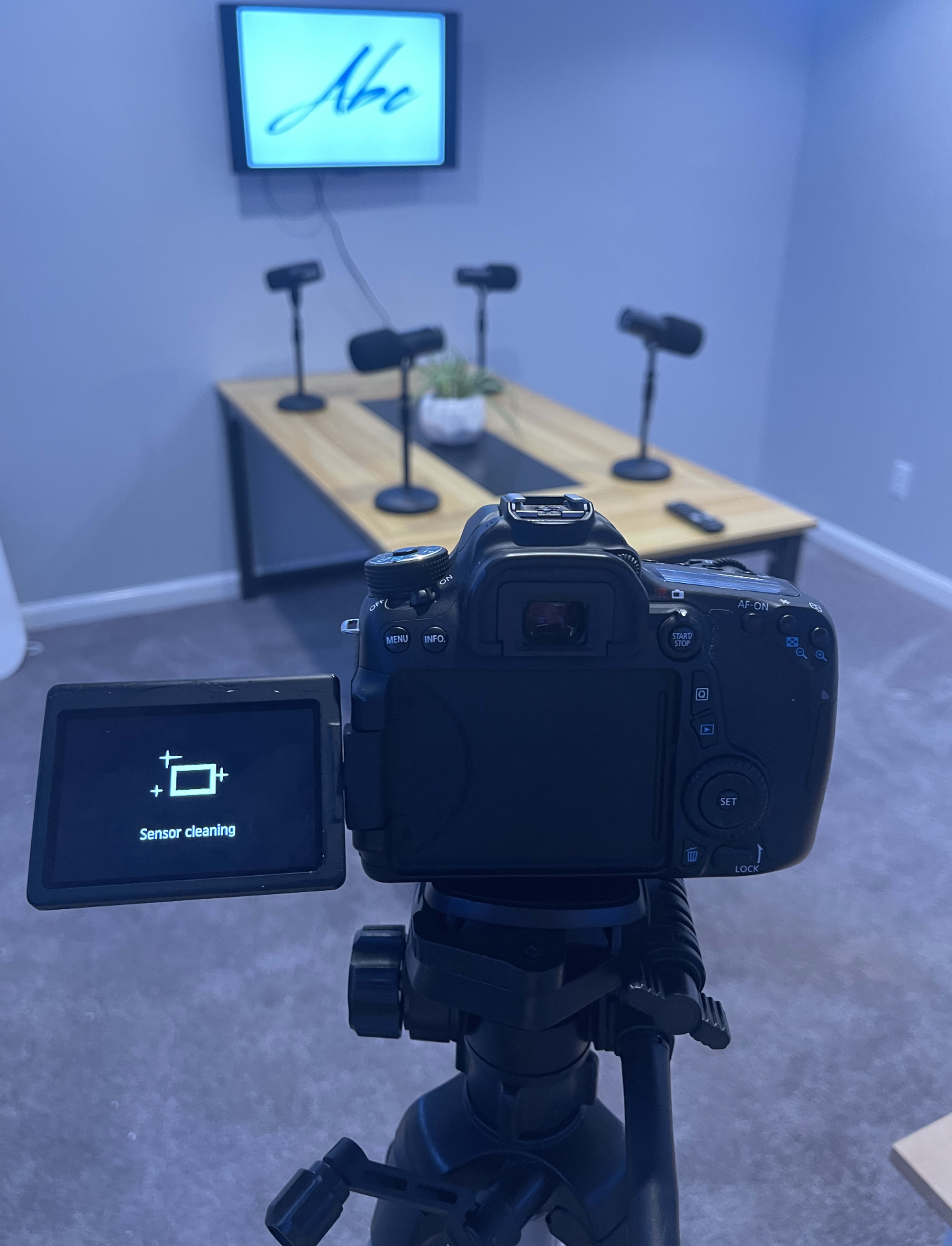 A camera on a tripod is positioned in front of a table with four microphones and a television displaying a logo in a well-lit room.