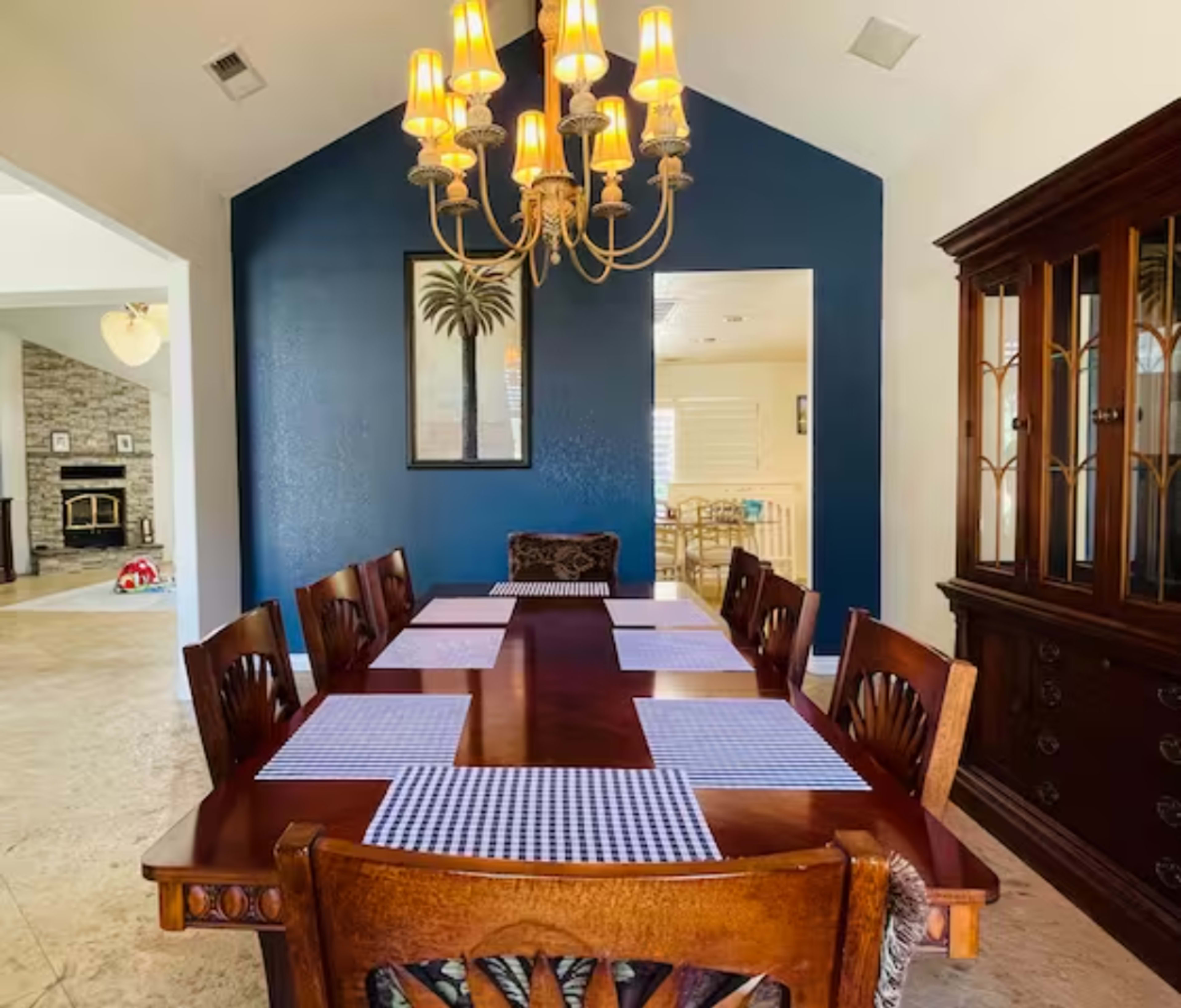 A dining room features a large wooden table set with placemats, surrounded by eight chairs, beneath a chandelier, with a blue accent wall and a glimpse of another room in the background.