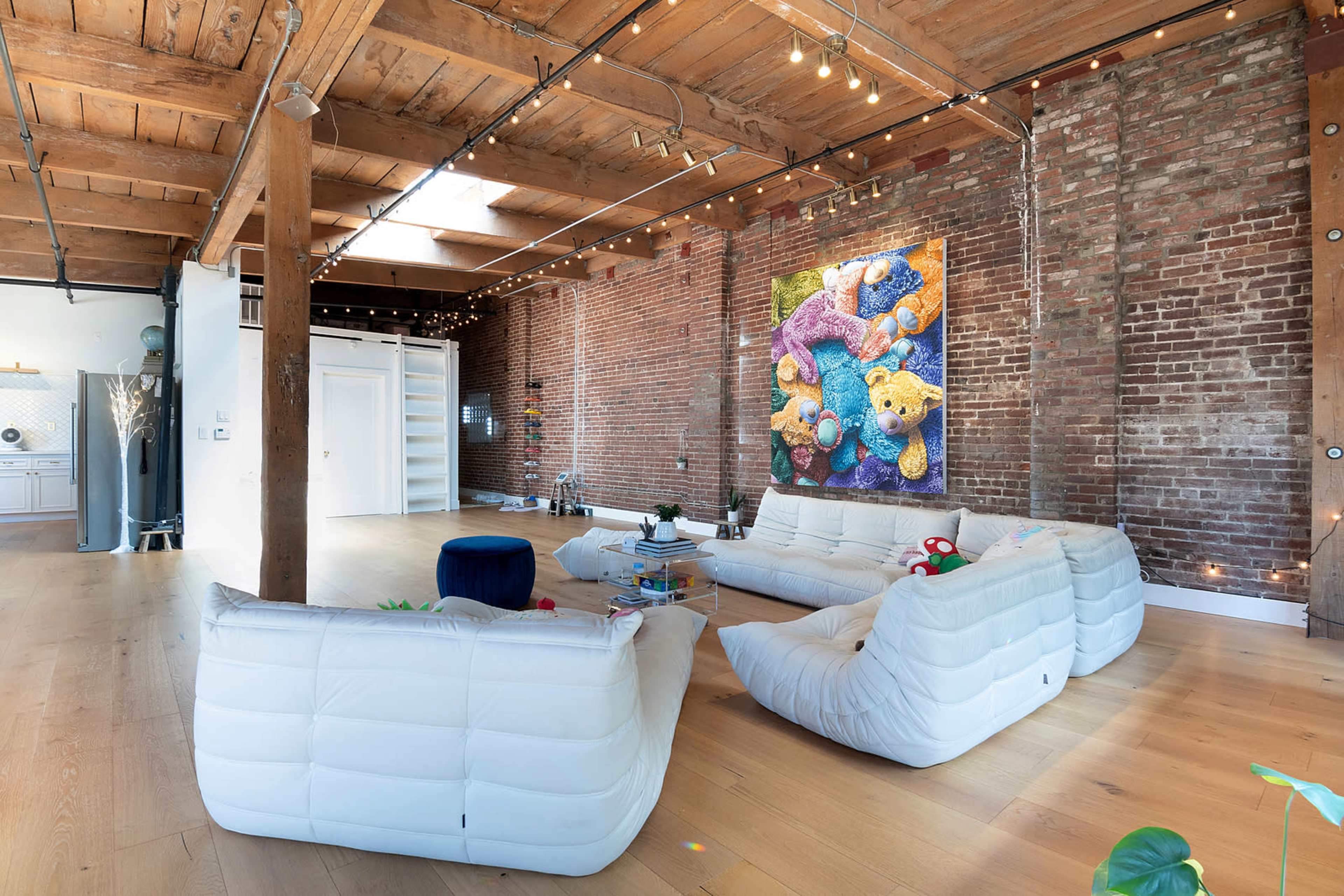 A spacious living area with exposed brick walls, wooden beams, and two white sofas arranged around a glass coffee table, featuring a colorful artwork on the wall.