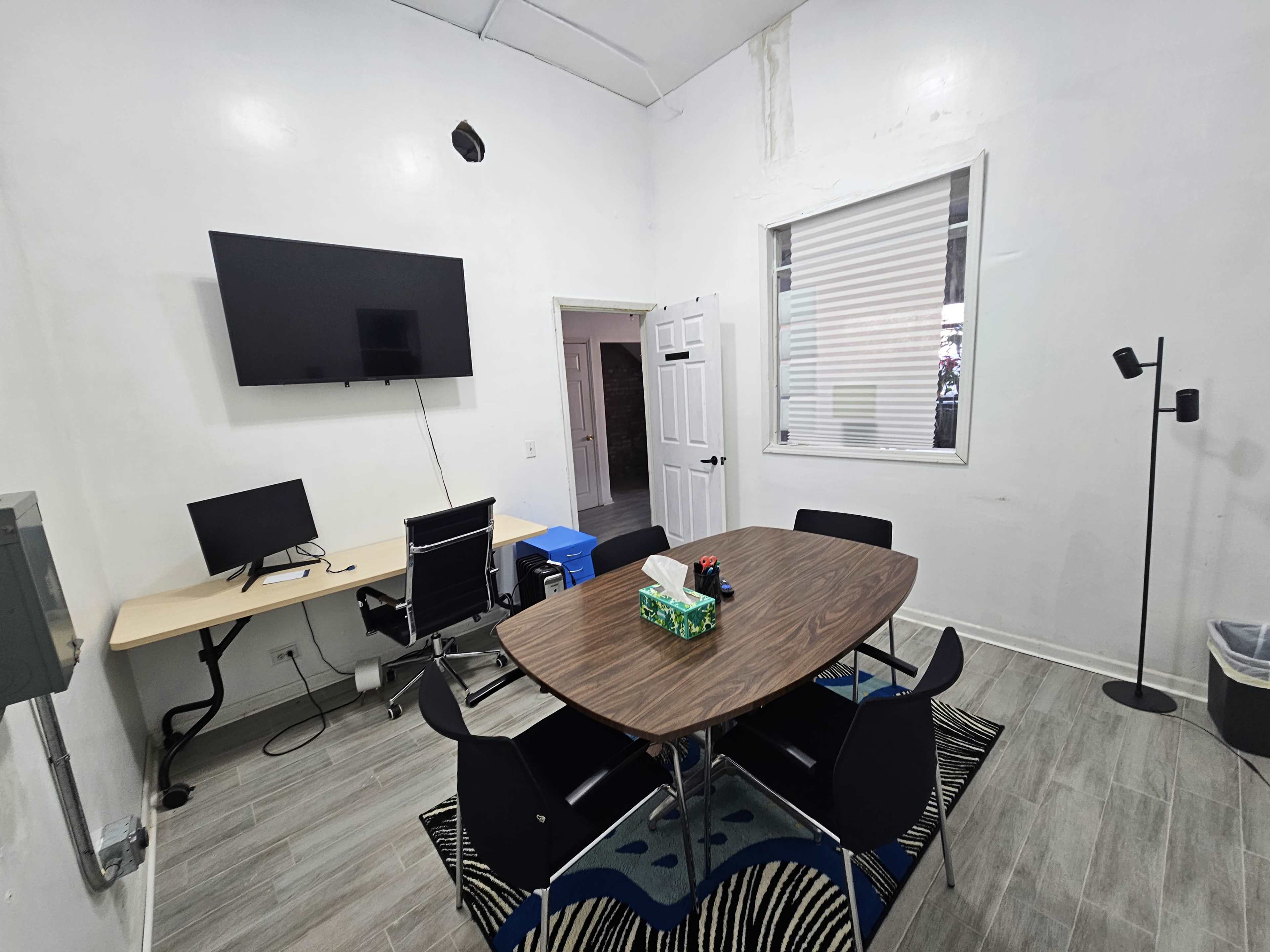 The image shows a simple office space featuring a table with chairs, a wall-mounted TV, and a desk with a computer setup.