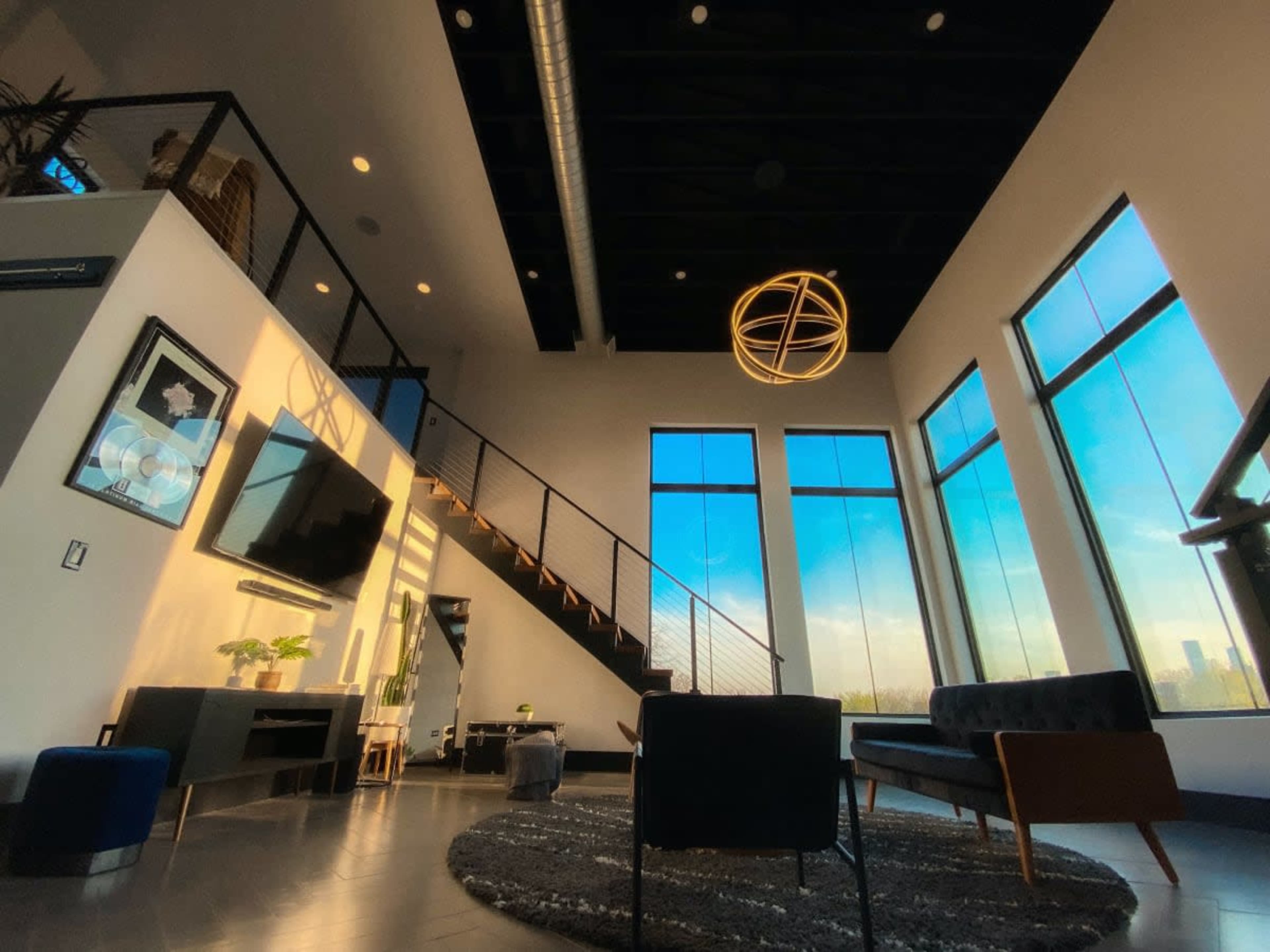 The image shows a spacious modern living room with large windows, a staircase, and contemporary furniture.