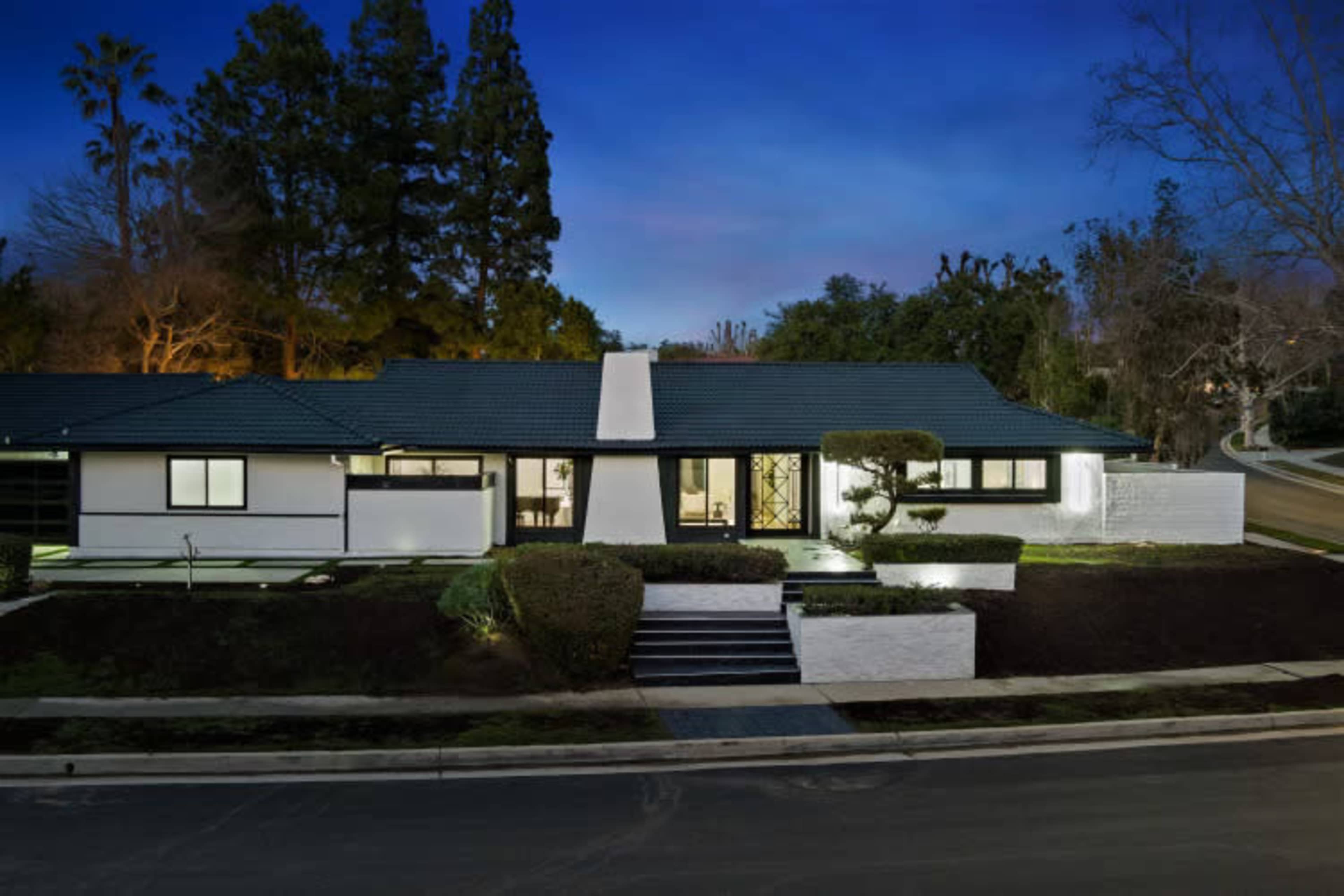 A modern, single-story house with a flat roof and landscaped front yard is seen at dusk.