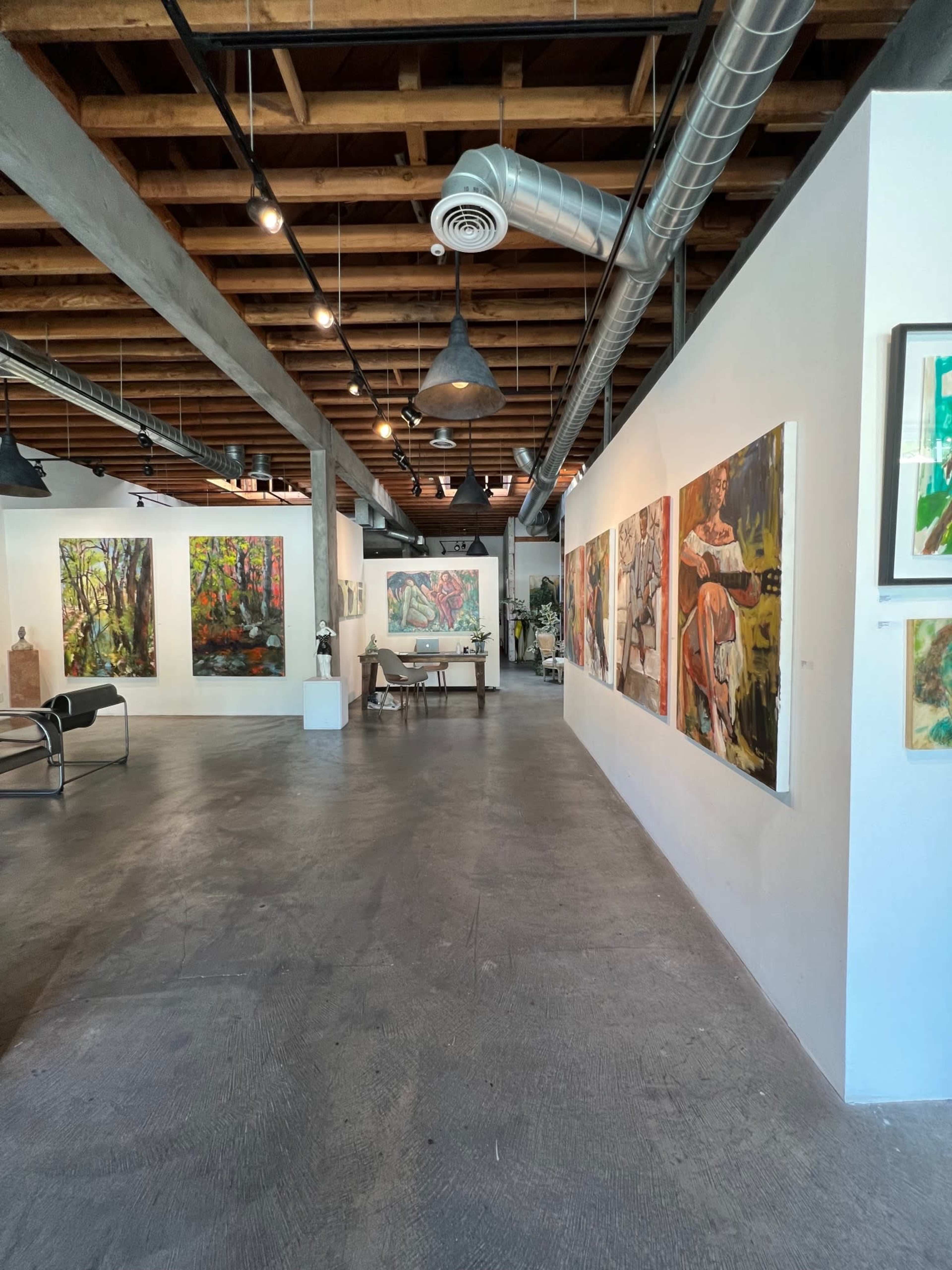 An art gallery with several colorful paintings displayed on white walls, featuring a central seating area and exposed wooden beams overhead.