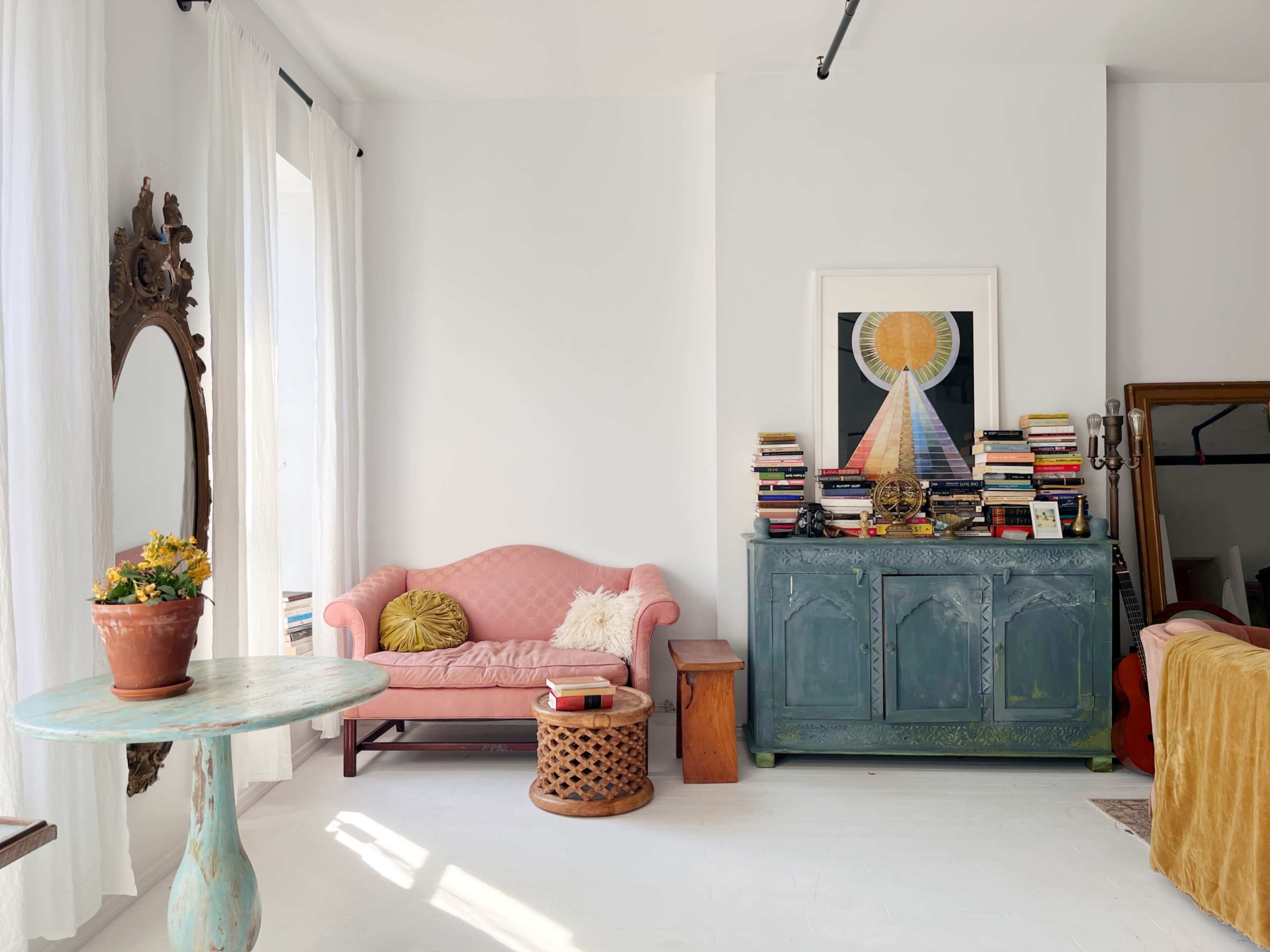 A pink sofa is positioned against a light-colored wall next to a green cabinet topped with books, with a round table and a potted plant in the room.