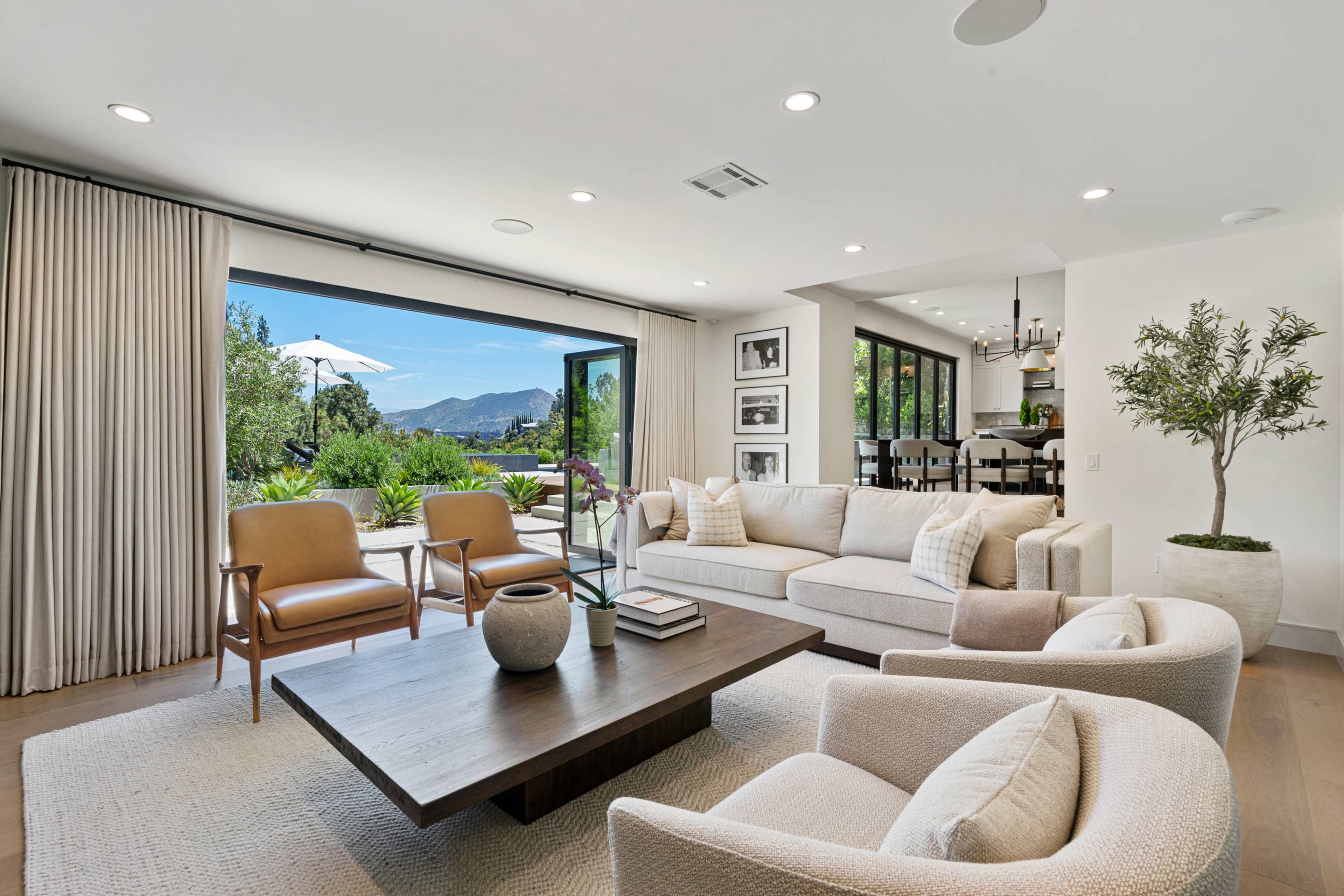 A modern living room with a light-colored sofa and chairs, a wooden coffee table, and large glass doors that open to a patio with a view of mountains and greenery.