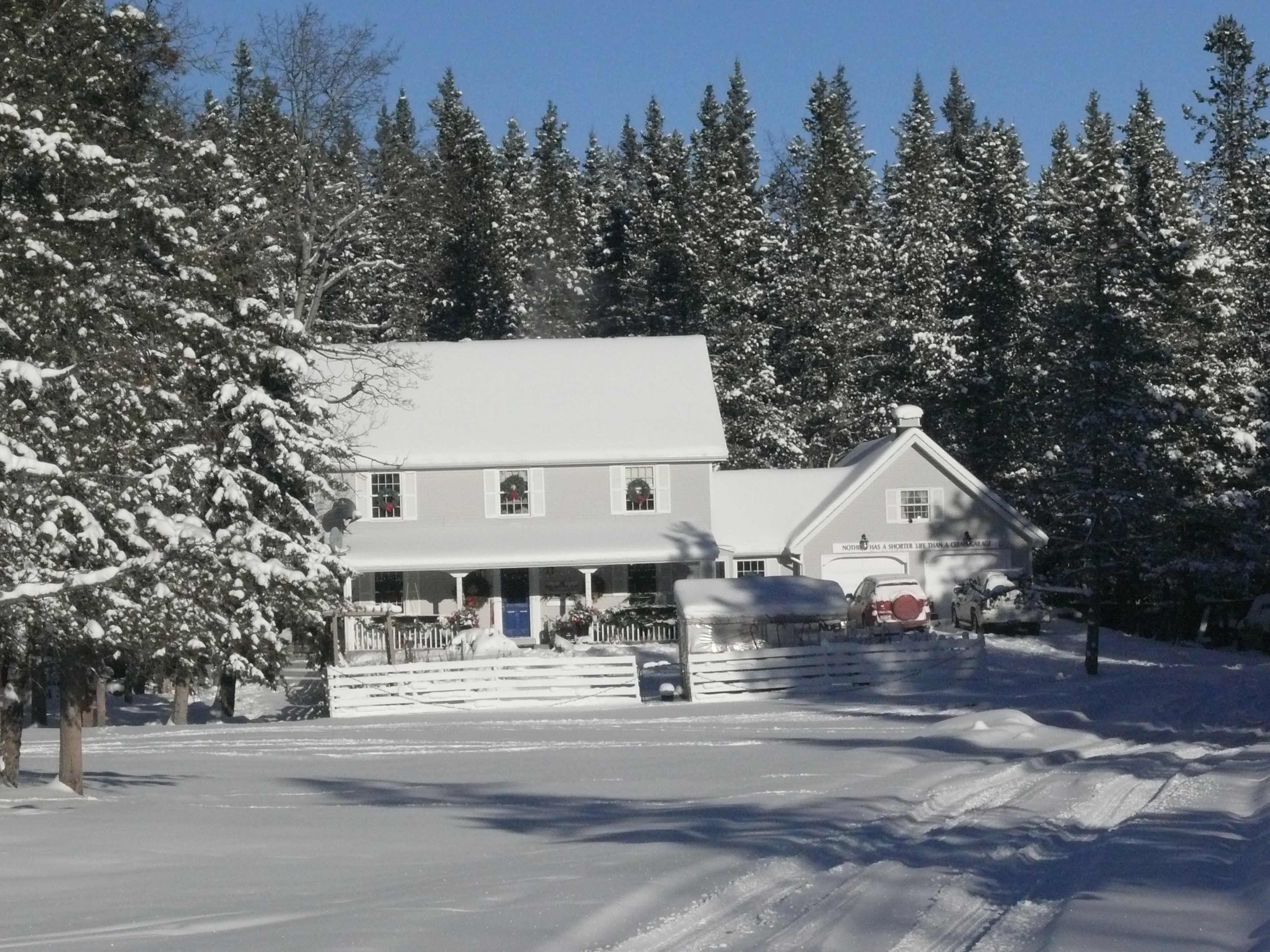 A two-story house with a sloped roof is surrounded by snow-covered trees and a white picket fence, with a clear blue sky above.