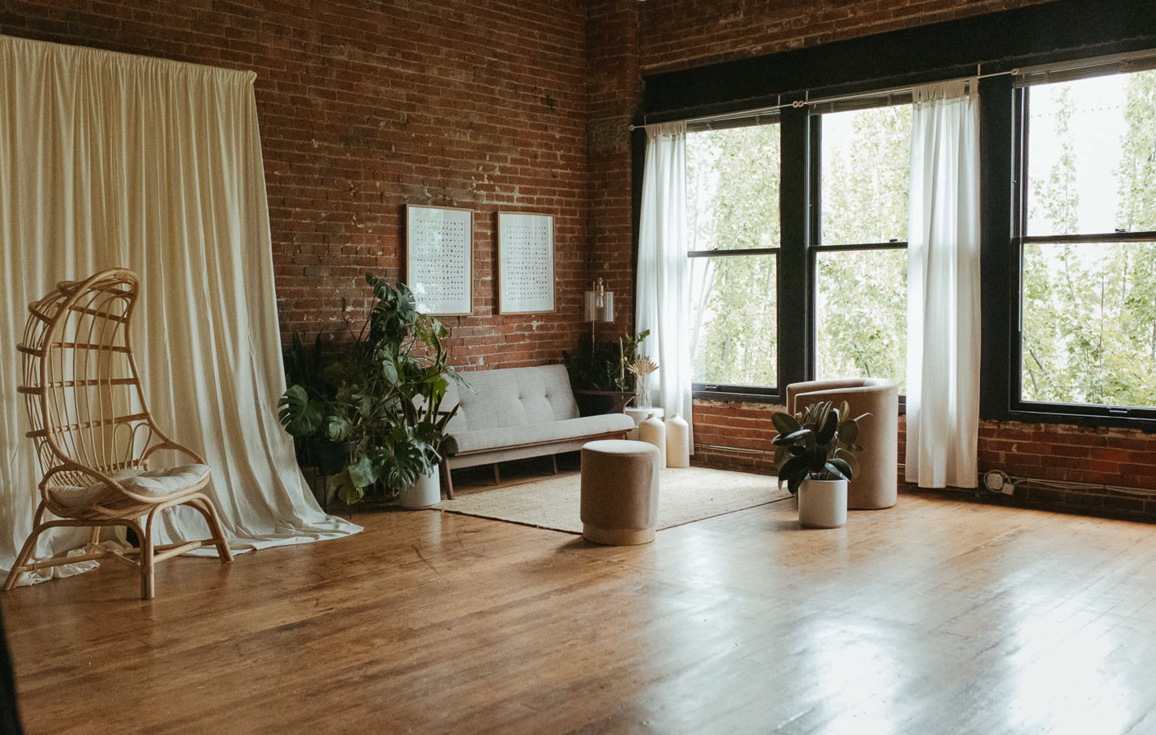The room features exposed brick walls, large windows with light curtains, a gray sofa, and various plants arranged with a rug on a wooden floor.