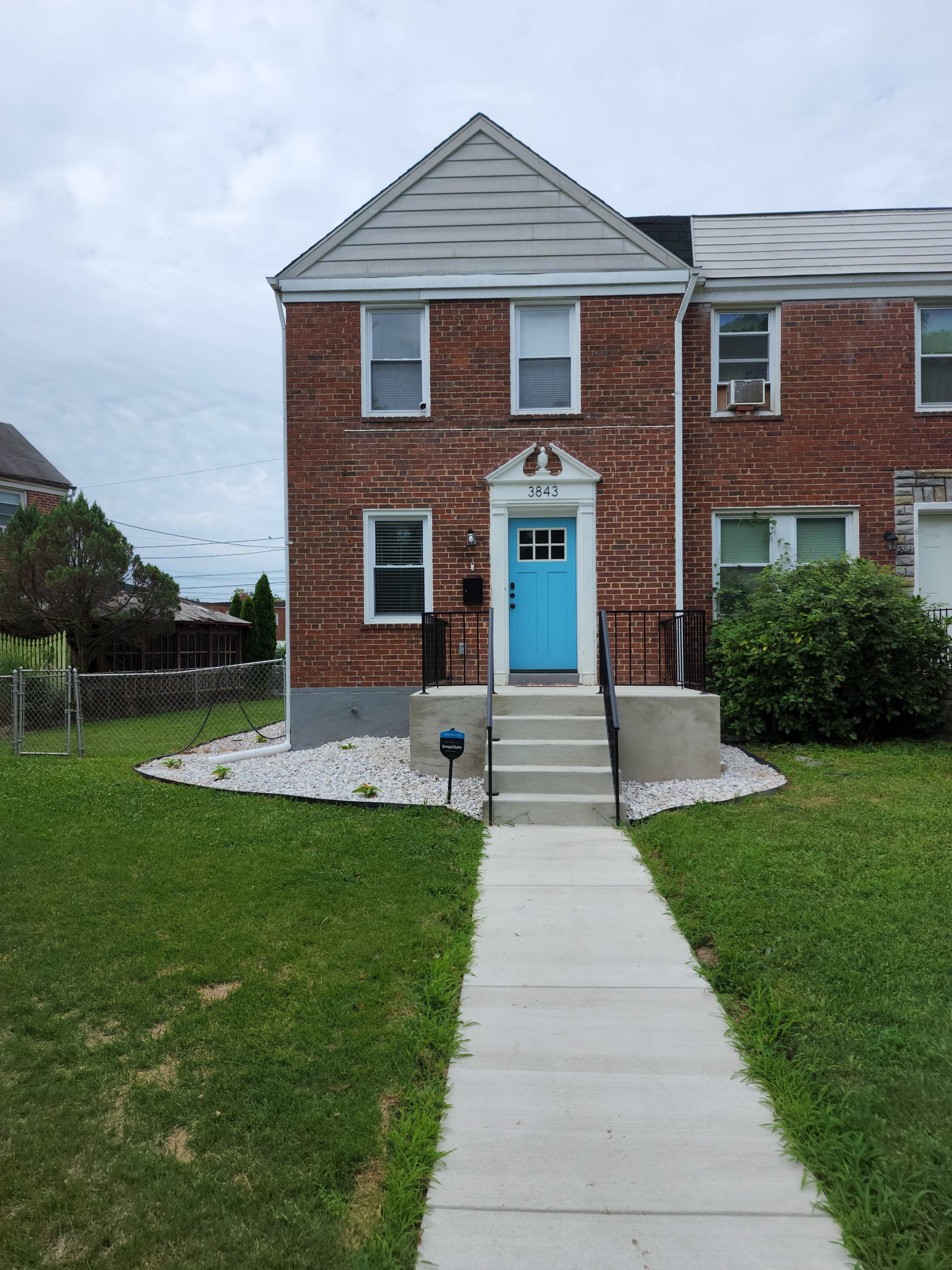 The image shows a brick front of a house with a turquoise door, a concrete walkway leading to it, and a grassy area in the foreground.
