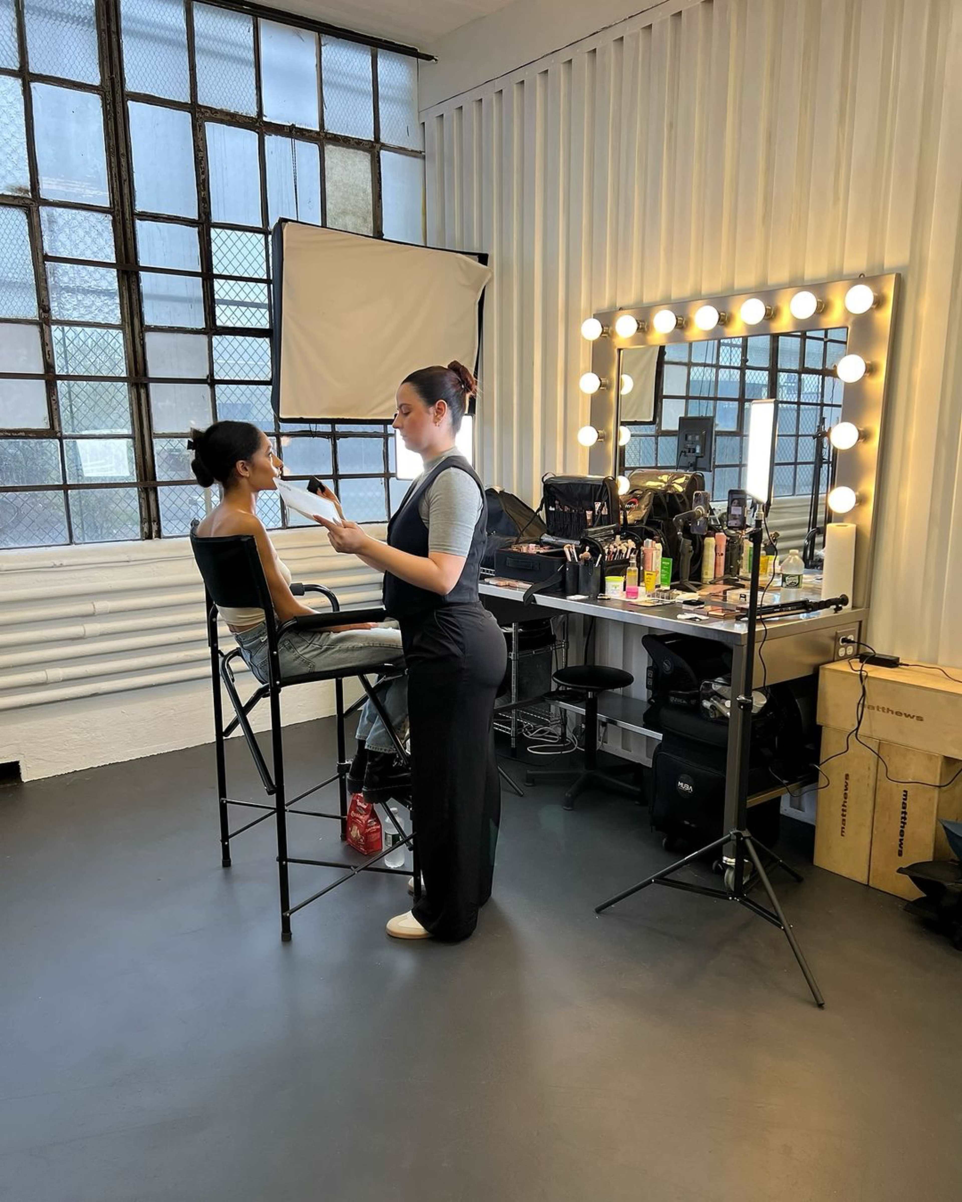 A makeup artist is applying makeup to a client seated on a high stool in a well-lit studio with a large mirror and various beauty products on the counter.