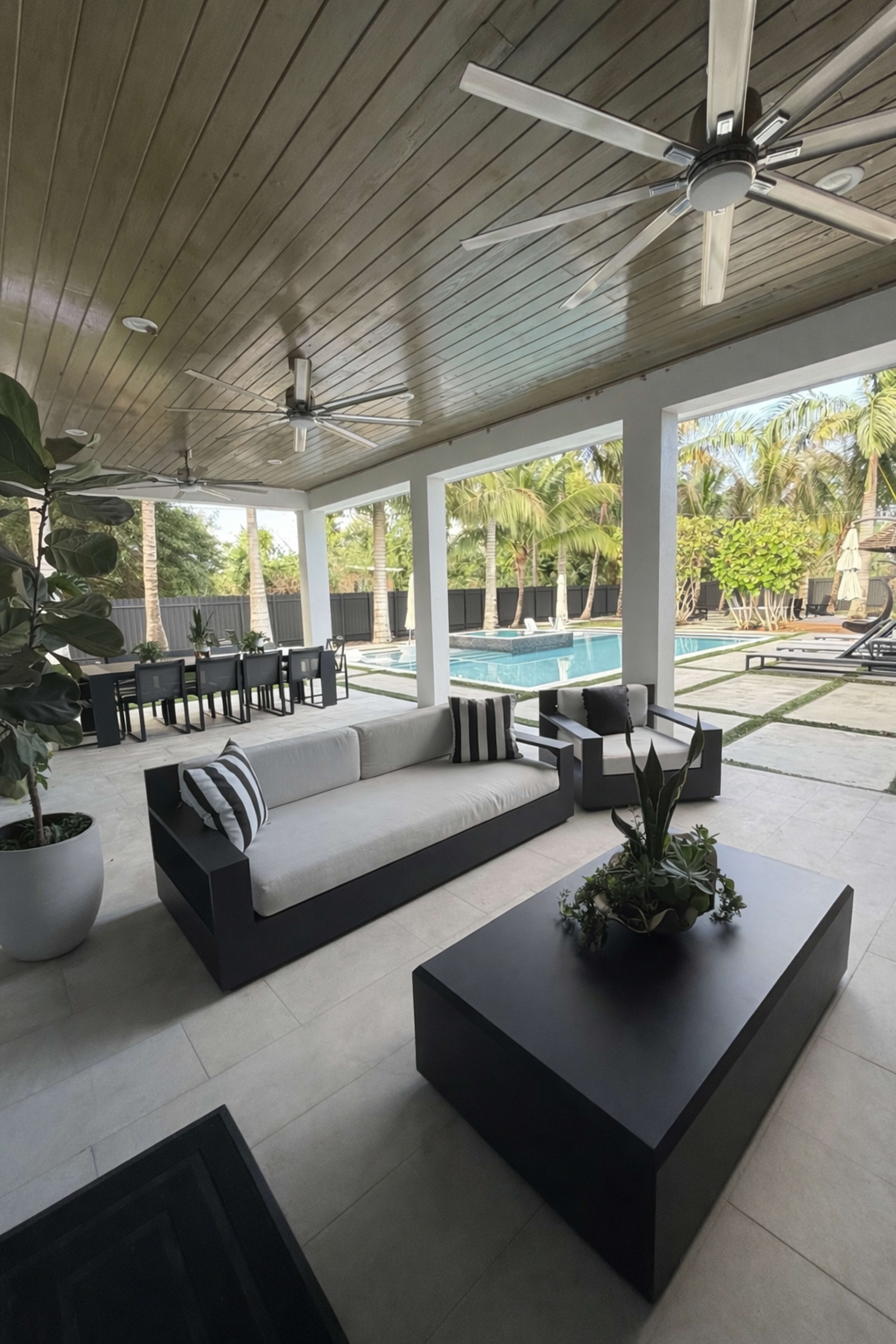 The image shows a covered outdoor living space featuring modern furniture, including a sectional sofa and a coffee table, with a view of a pool and landscaped backyard.