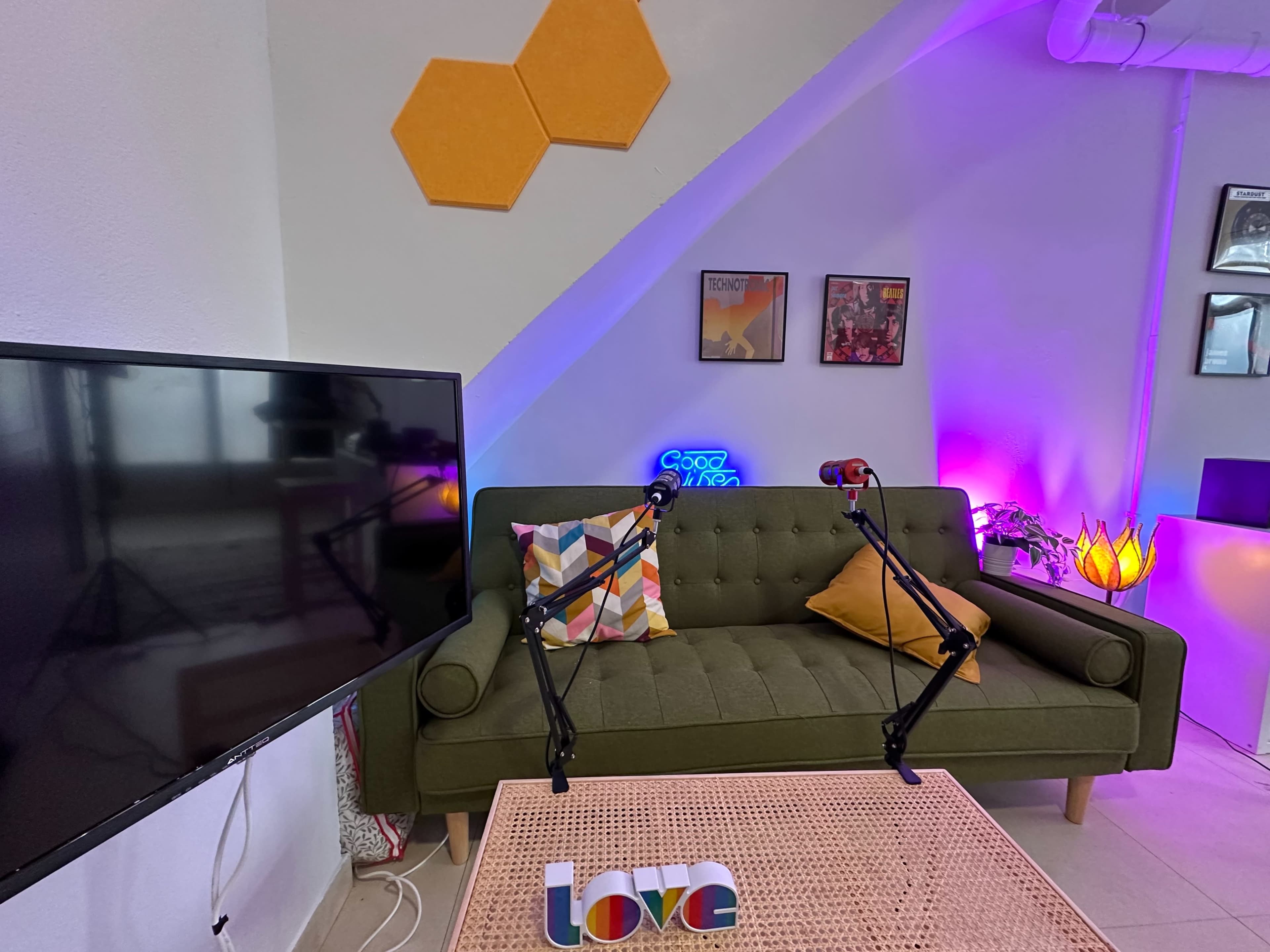 A green sofa with decorative pillows, a television, and a colorful coffee table in a room with artistic wall decor and purple lighting.