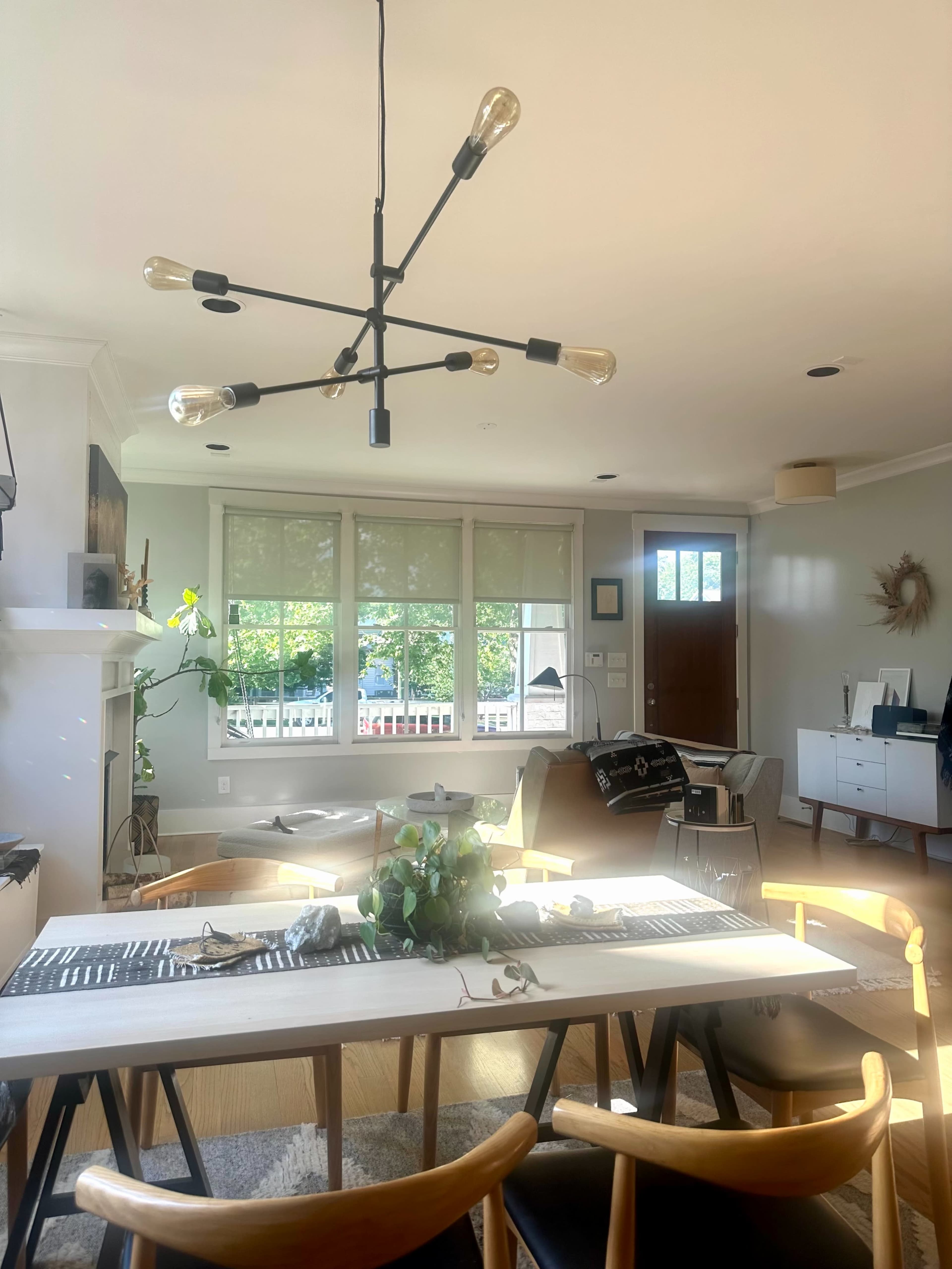 A modern living space features a light fixture above a dining table, surrounded by chairs, with windows allowing natural light and a view of the outdoors.