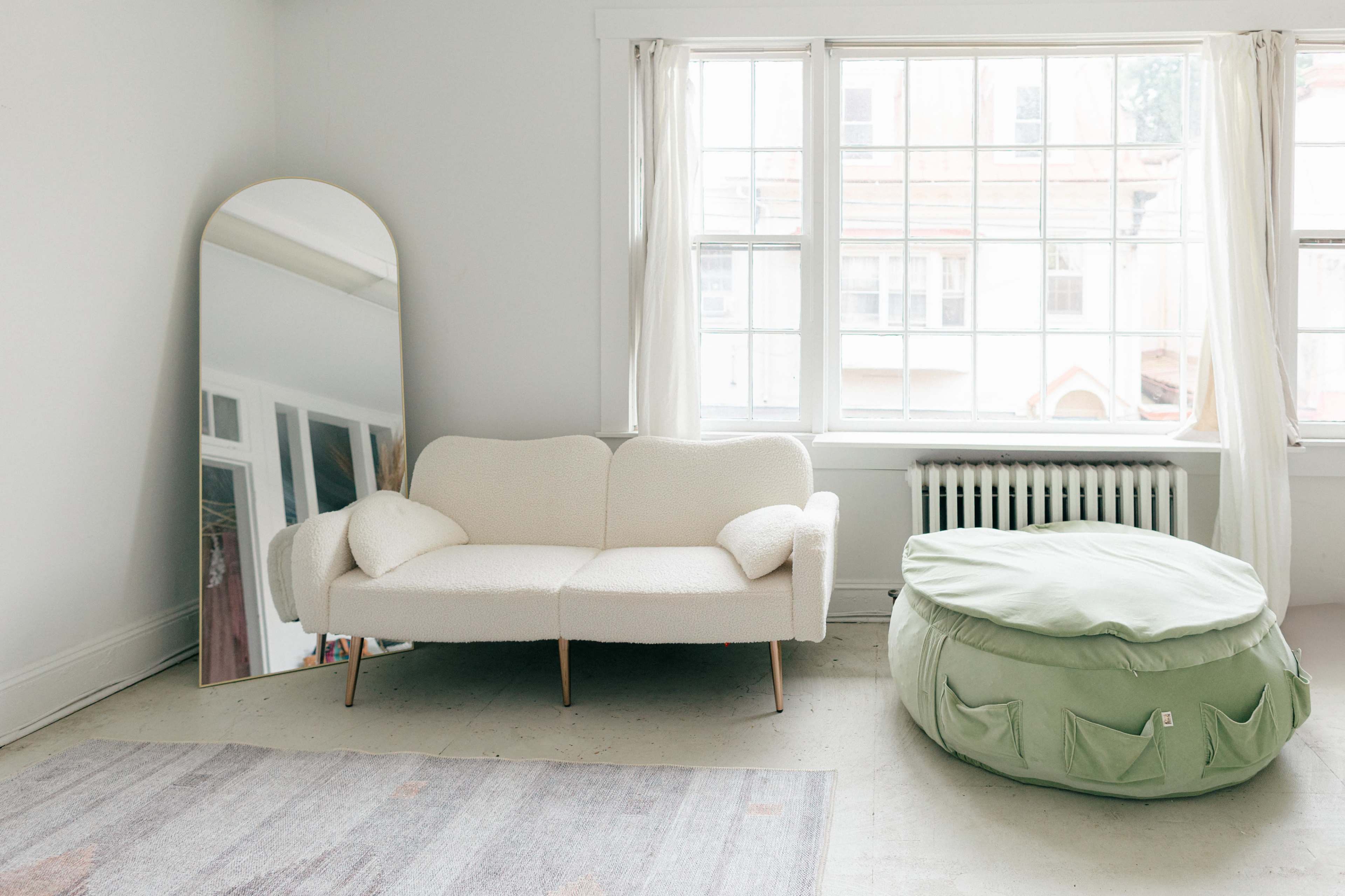 A light-filled room features a plush white couch, a large mirror, and a green pouf, with a radiator beneath a window.