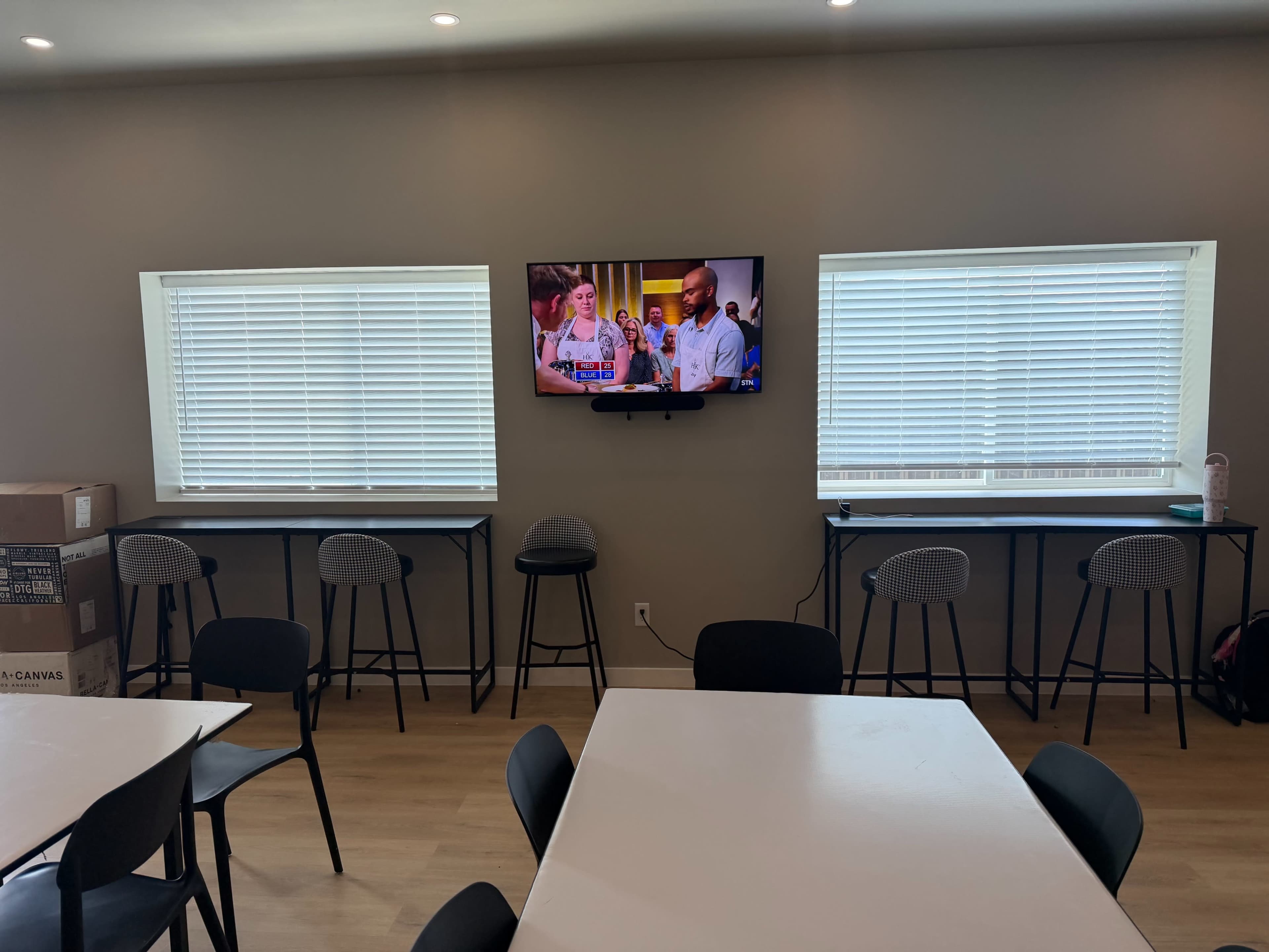 The image shows a room with a table and several black chairs, featuring a television mounted on the wall between two windows with blinds, and bar-style seating beneath the windows.