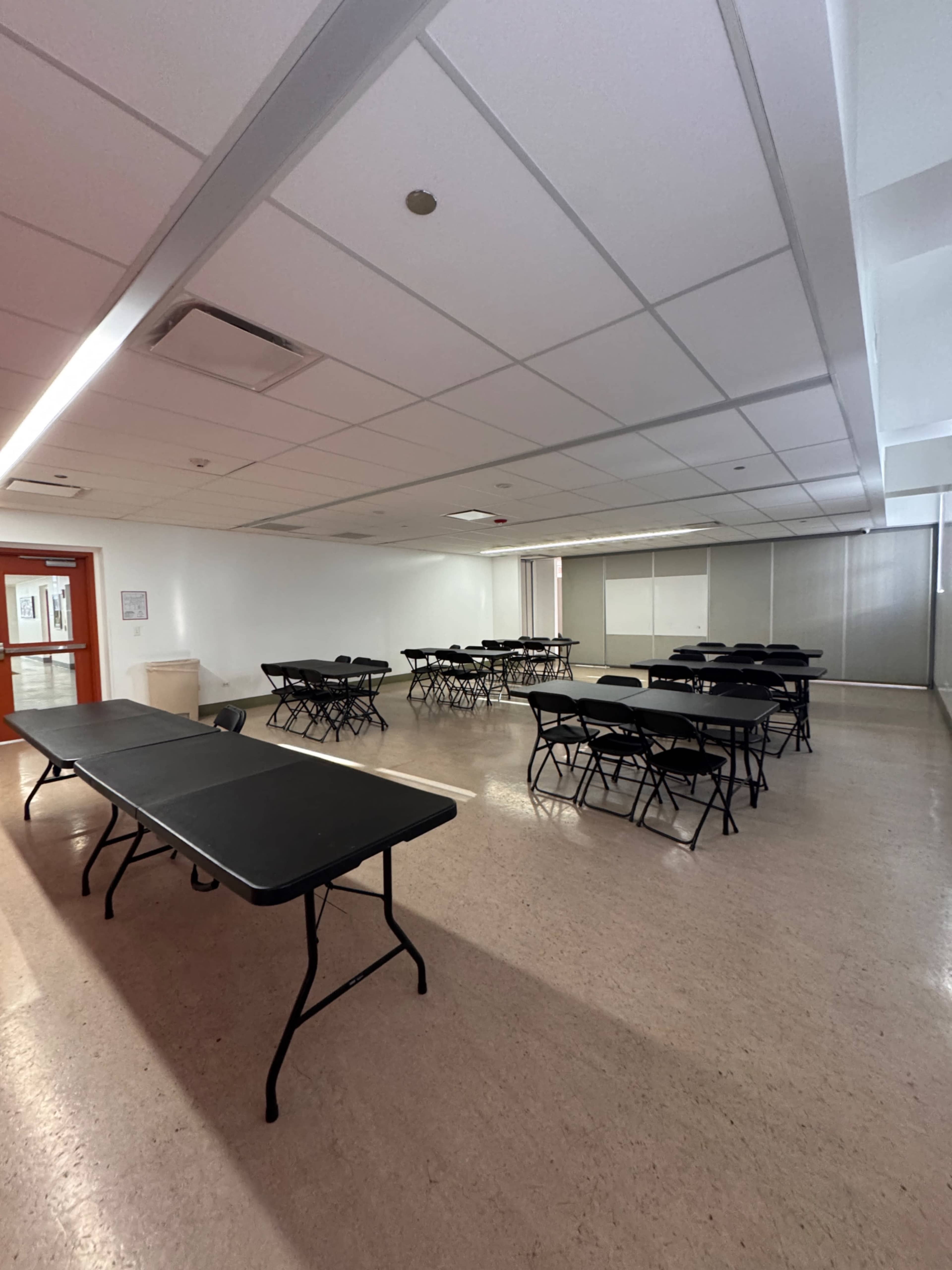 The image shows a spacious room arranged with several black folding tables and chairs, all positioned evenly throughout the area.
