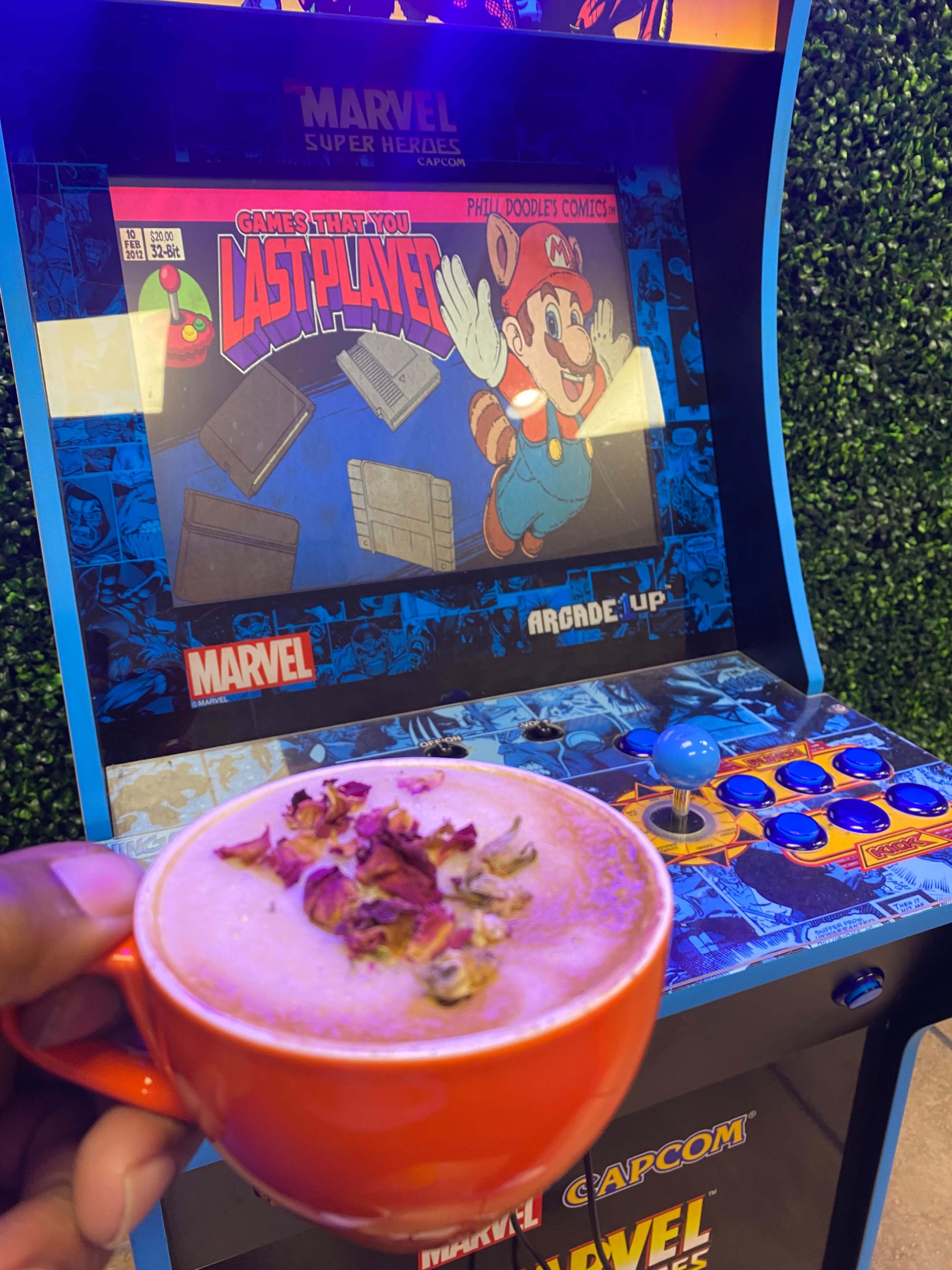 A person holds a bright orange cup of coffee topped with rose petals in front of an arcade cabinet featuring a Marvel video game.
