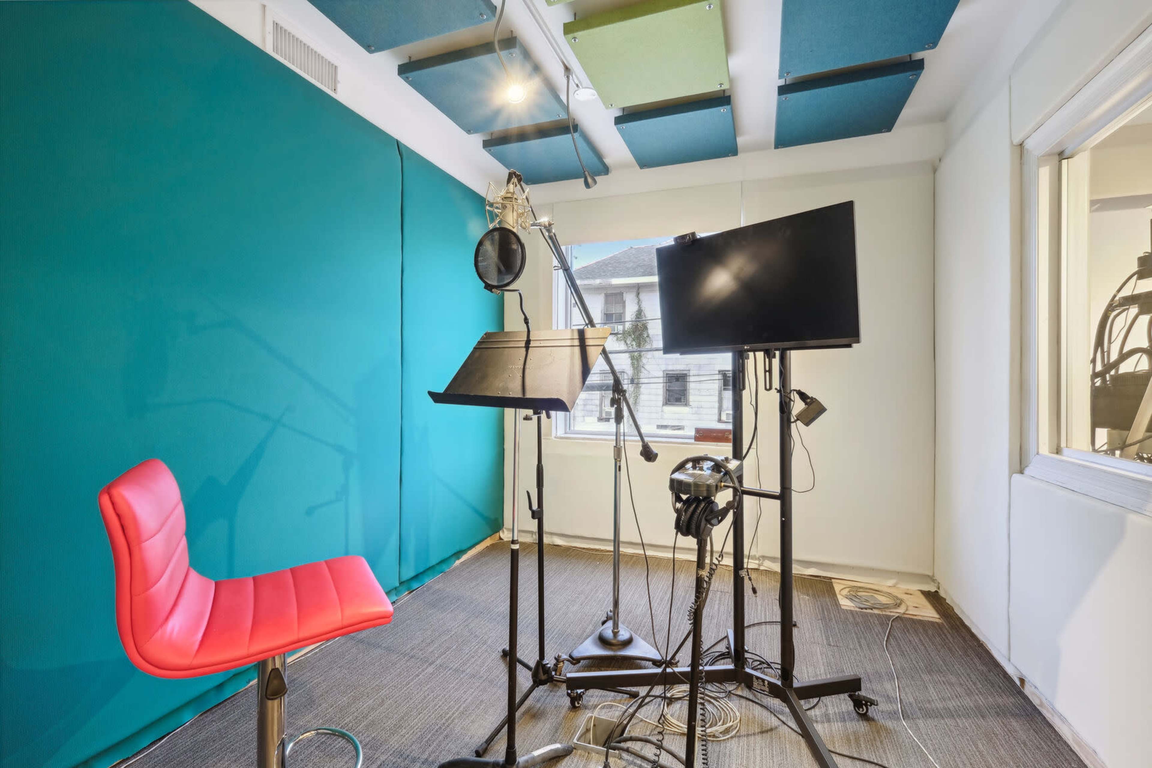 The image shows a small recording studio with a red chair, a microphone setup, a music stand, and a monitor.