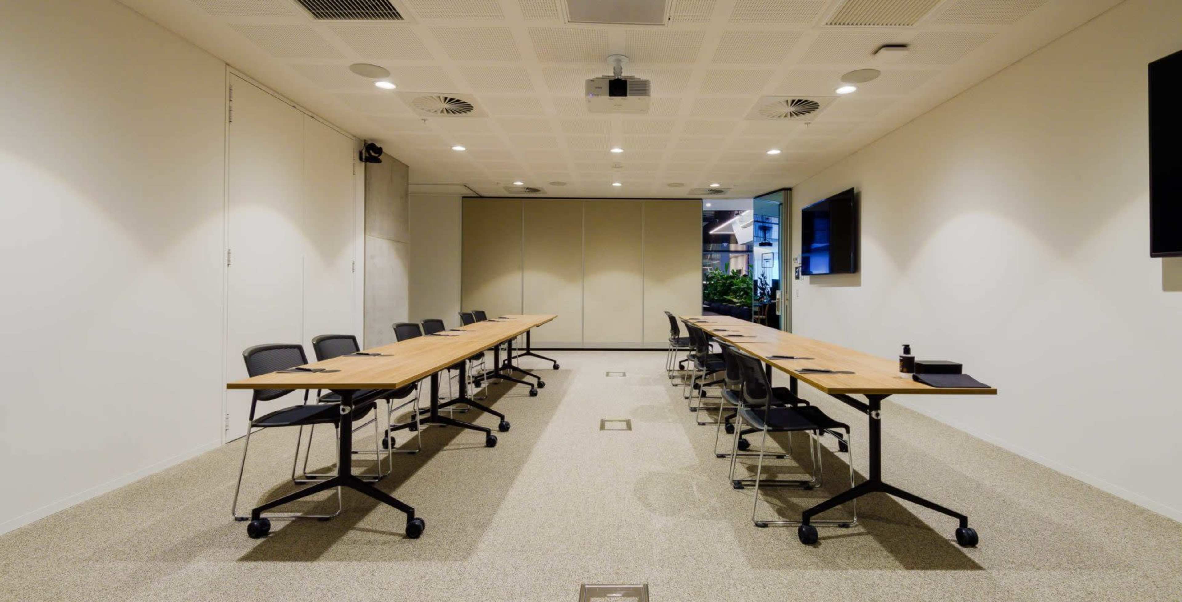 A modern conference room features a long wooden table with black chairs arranged on either side and two screens mounted on the walls.