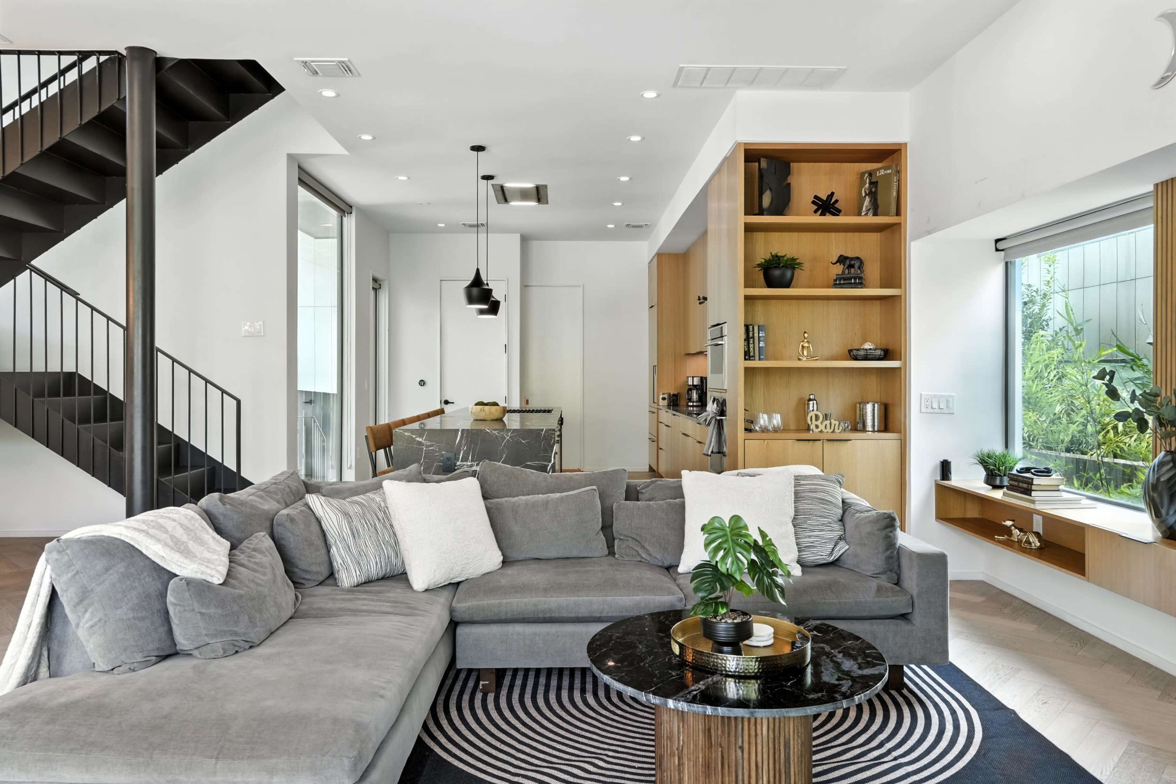 The image shows a modern living room with a large gray sectional sofa, a circular coffee table, a staircase, and an open kitchen area with wooden shelves.