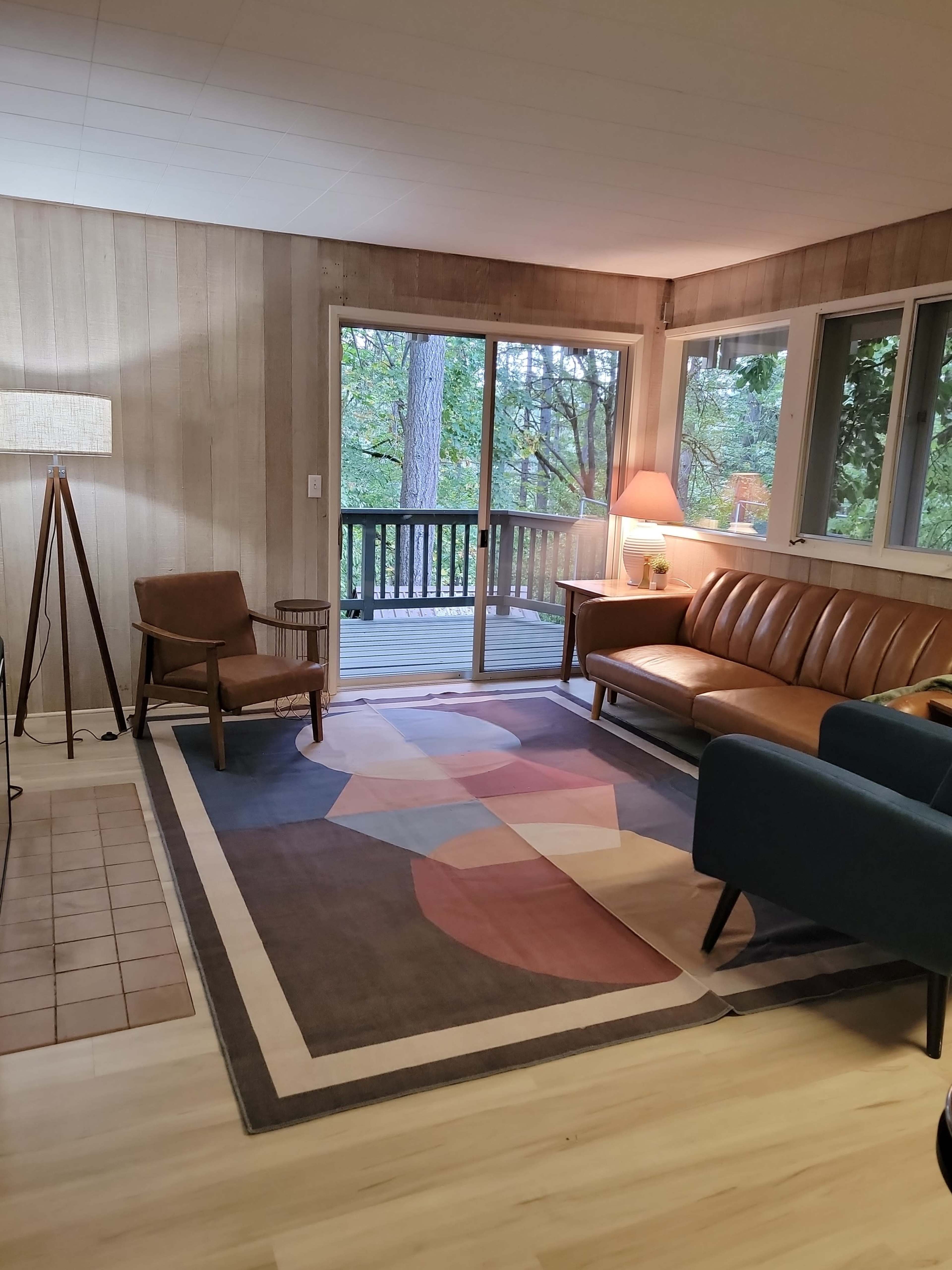 The image shows a cozy living room with a brown leather sofa, a blue chair, a wooden coffee table, and large windows overlooking a wooded area.