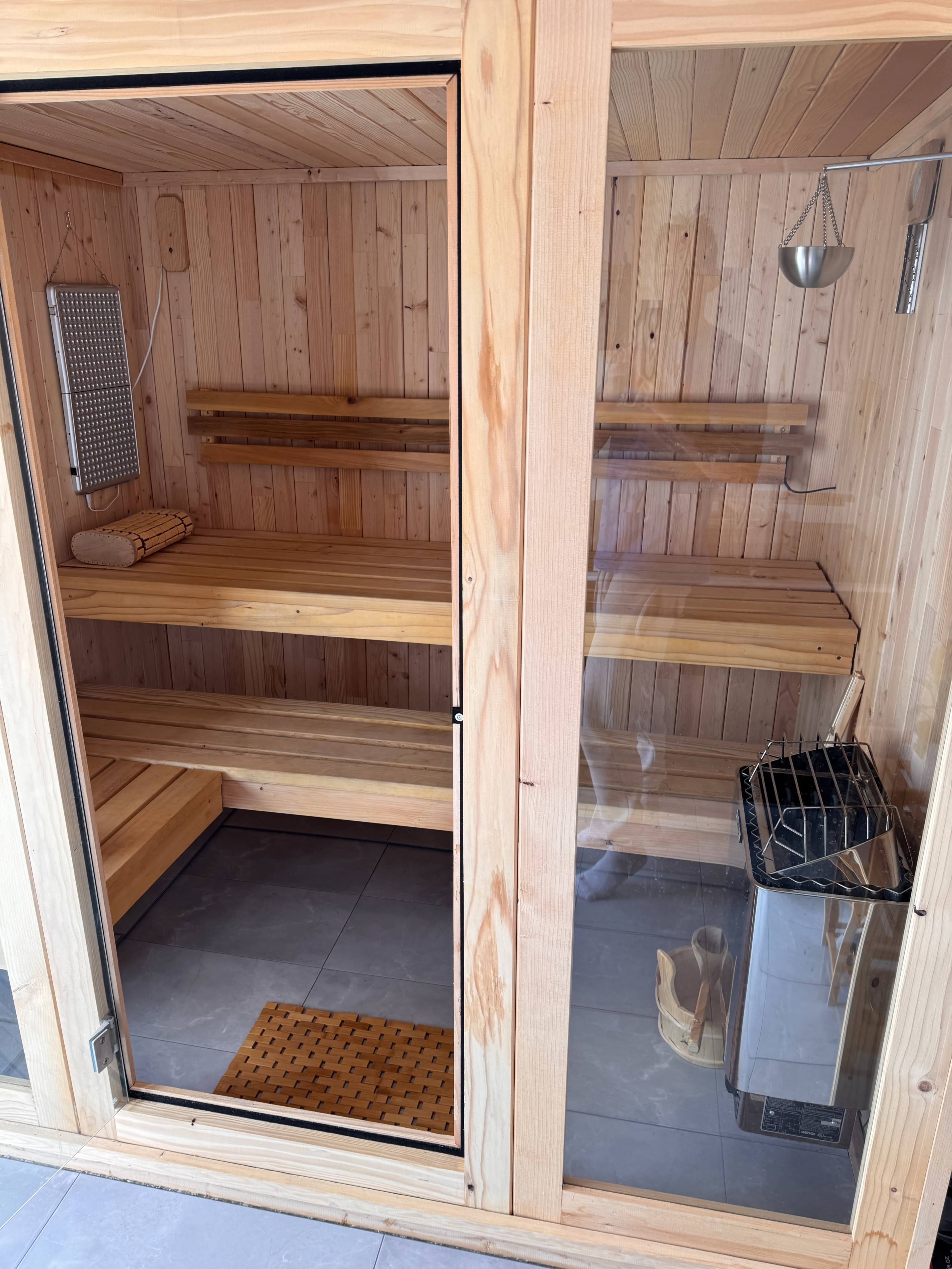 The image shows a wooden sauna room with benches, a heater, and a glass door.